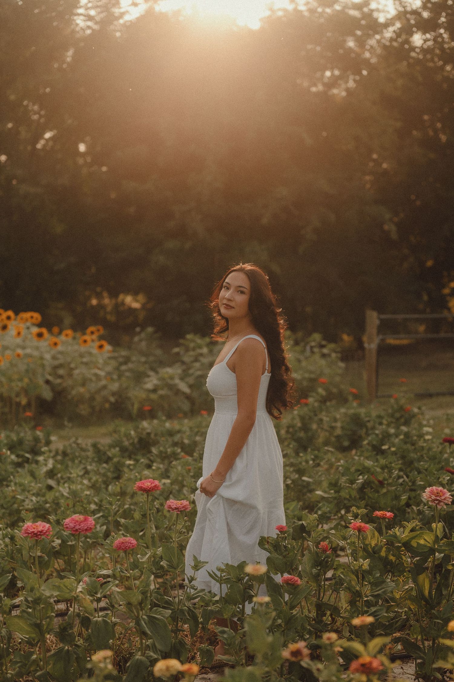 Woman in white dress standing in flower garden at golden hour sunset with sunflowers and pink blooms.