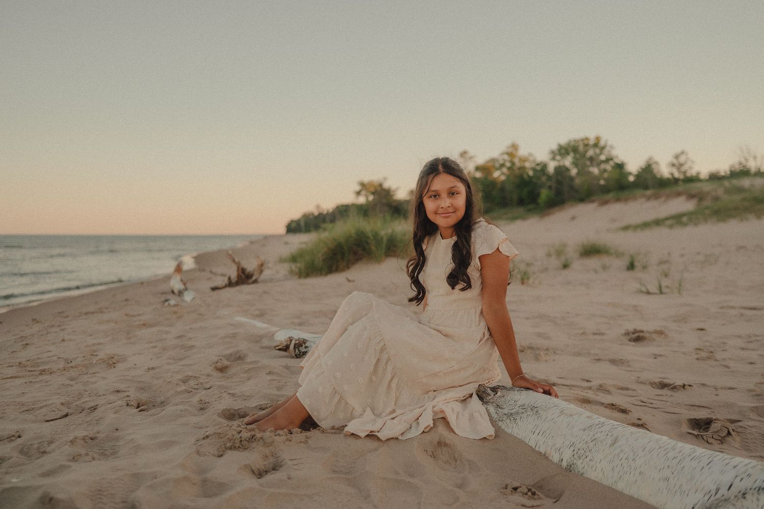 A young girl in a flowing beige dress sits on driftwood at a sandy beach during golden hour sunset.