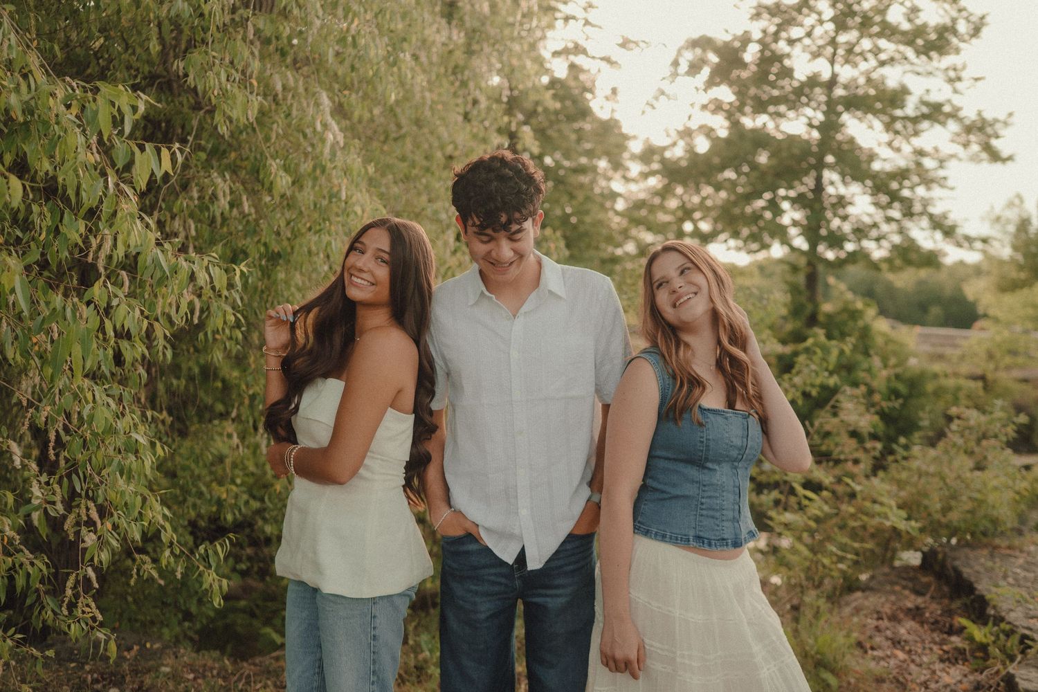 Three young adults smiling and posing together outdoors among lush green trees in warm golden light.