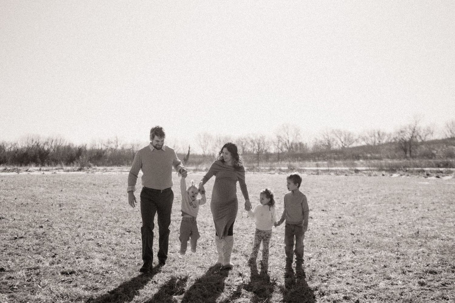 A family of four walking together through a field, viewed from behind in black and white.