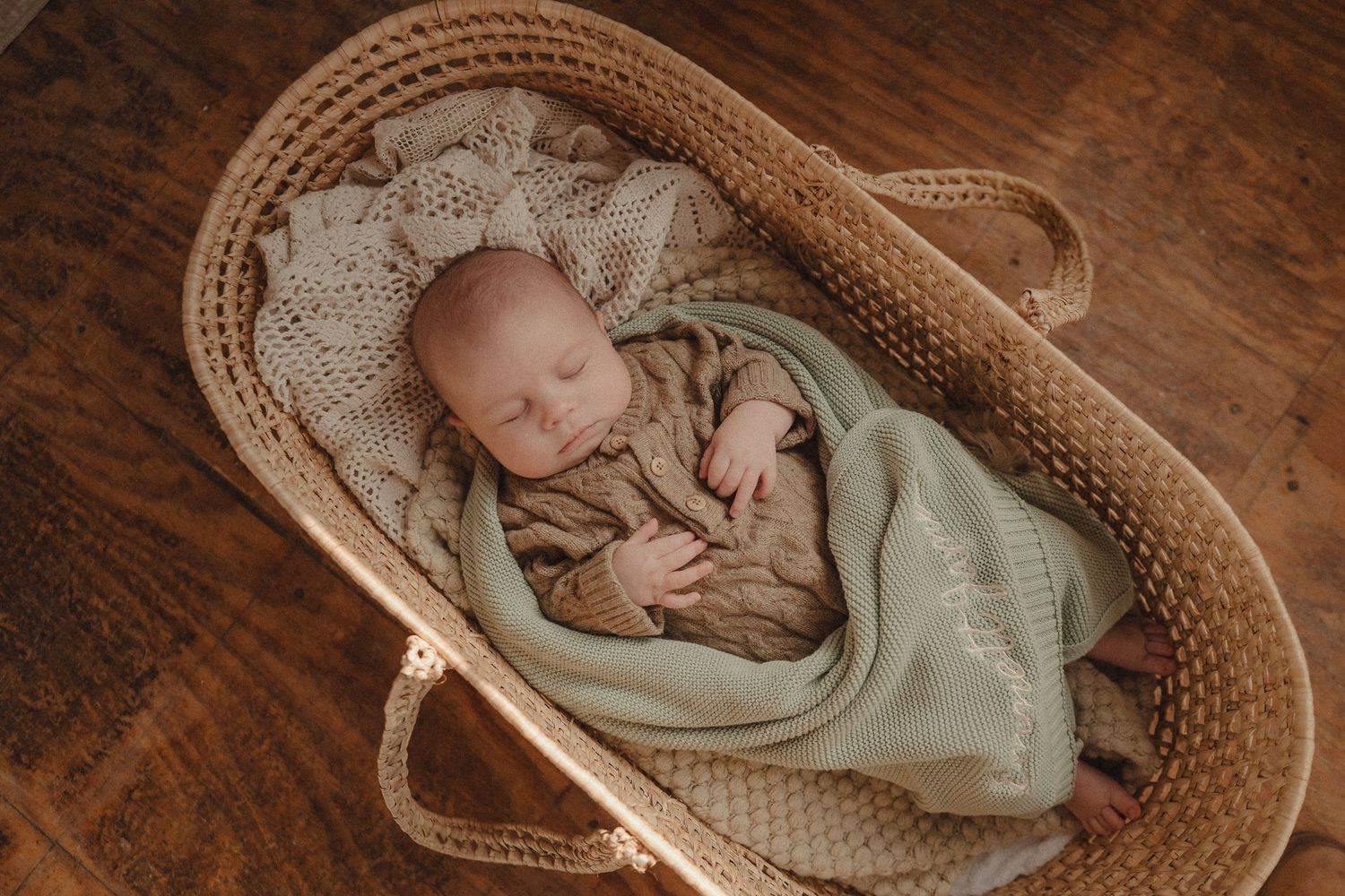 Baby resting in vintage woven basket on wooden floor with mint green swaddle.