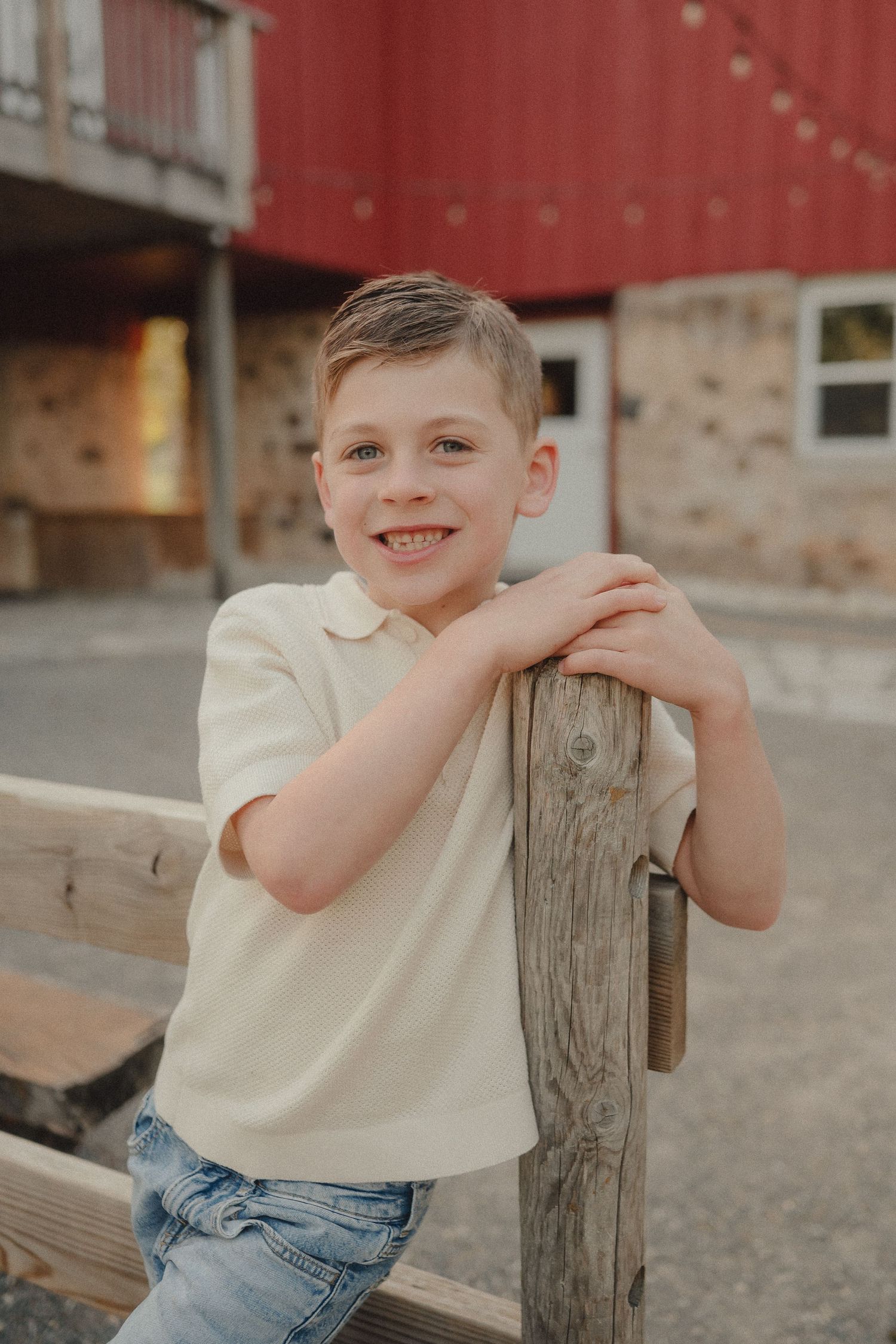 A cheerful young boy in a cream polo shirt leaning on a wooden fence post near a red barn.