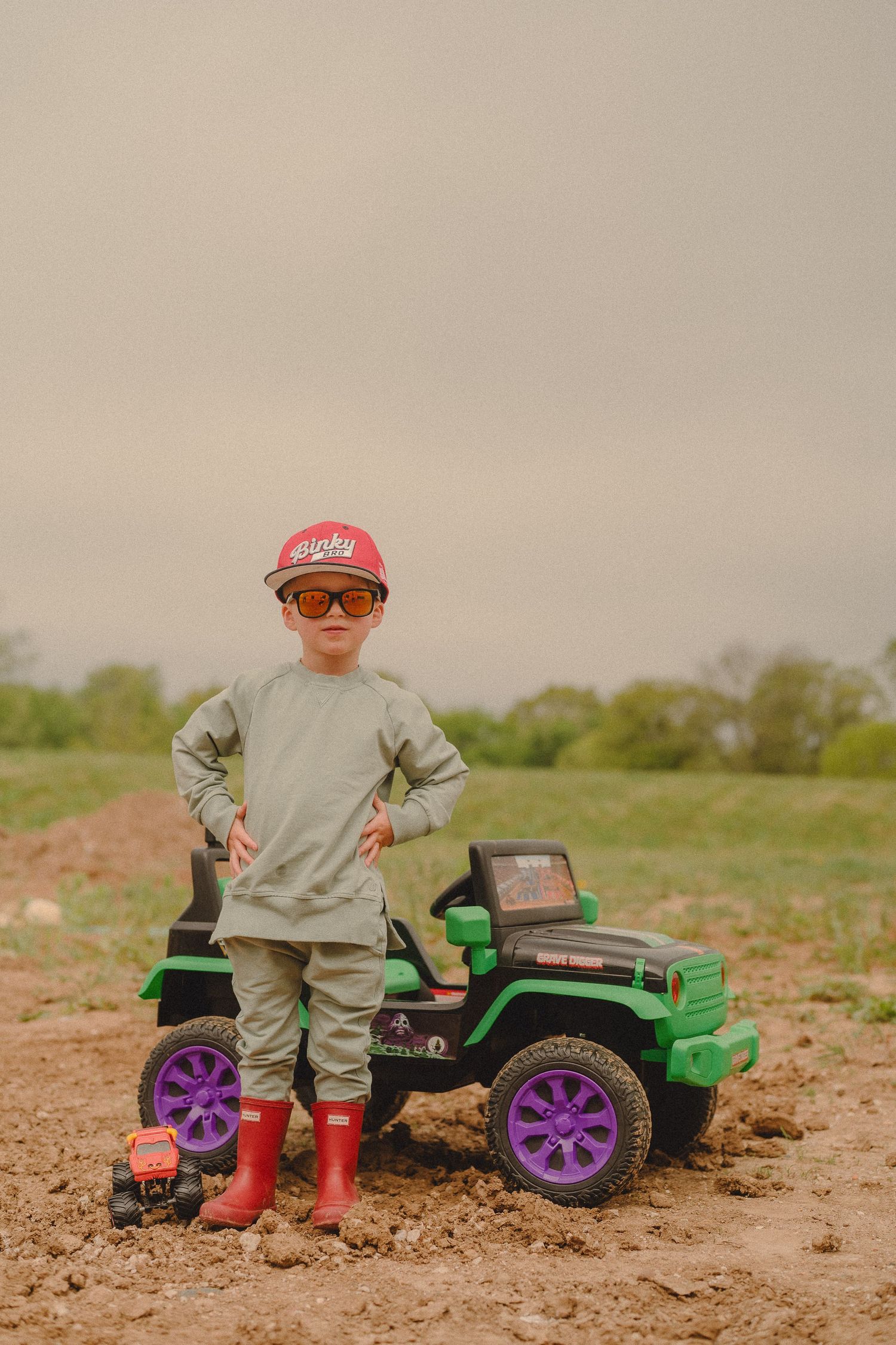Cool boy in red cap and sunglasses poses beside green Monster Truck ride-on toy in mud.