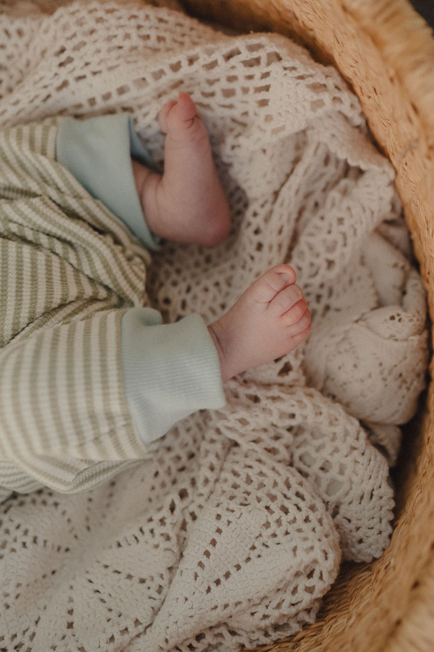 Close-up of newborn baby feet in green striped onesie resting in wicker basket with knit blanket.