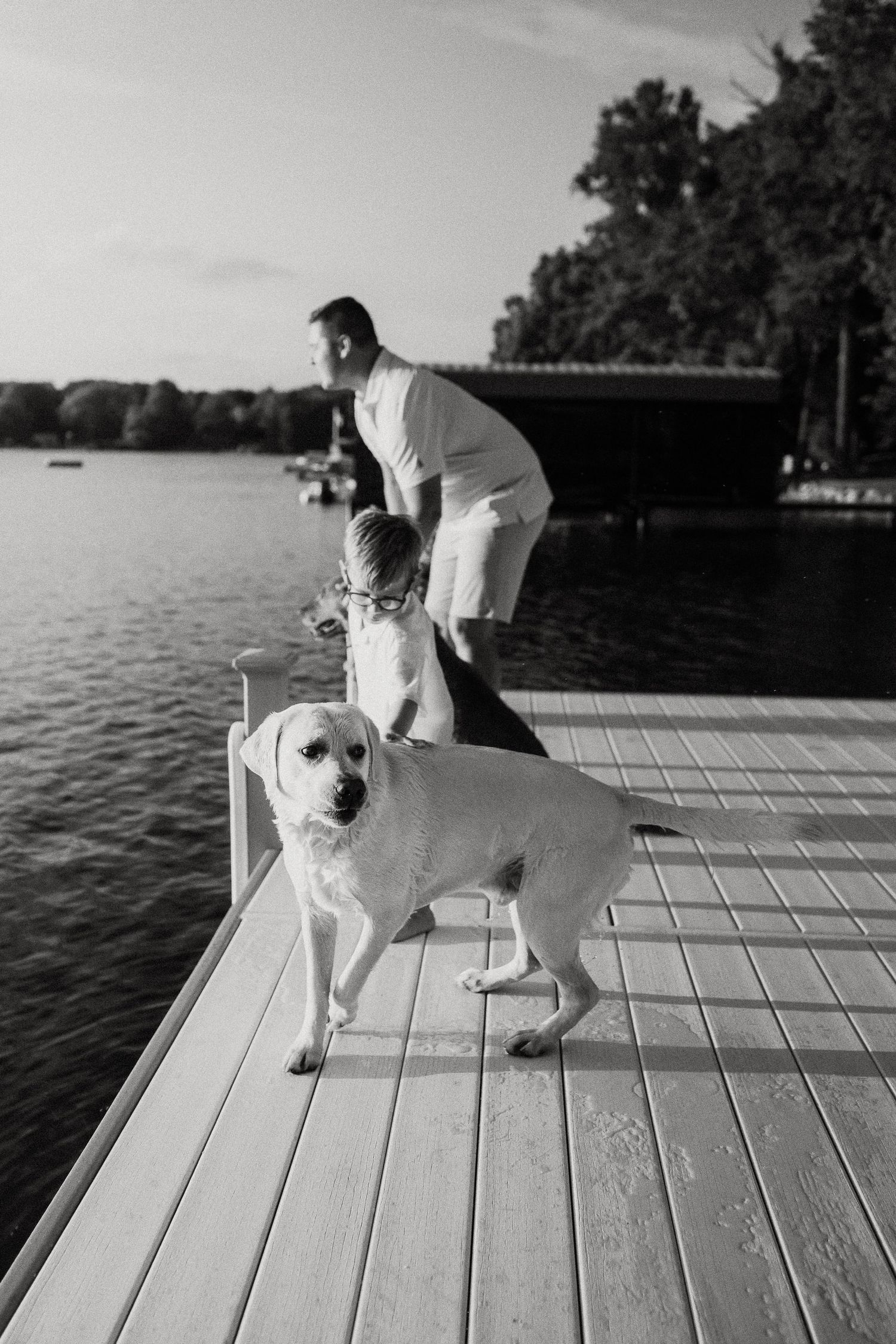 Man and child stand on a lakeside dock with a white Labrador dog in a black and white photo.