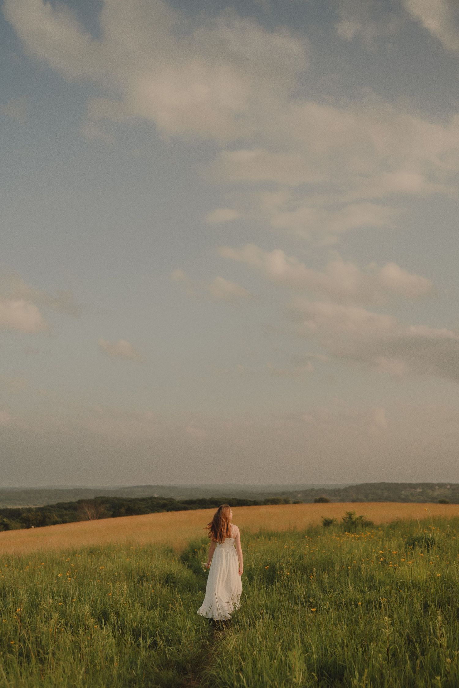 Woman in white dress walking along a path through a green field toward the horizon under a cloudy sky.