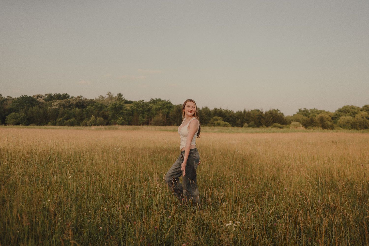 A woman stands smiling in a vast golden meadow with lush green trees in the background during golden hour.