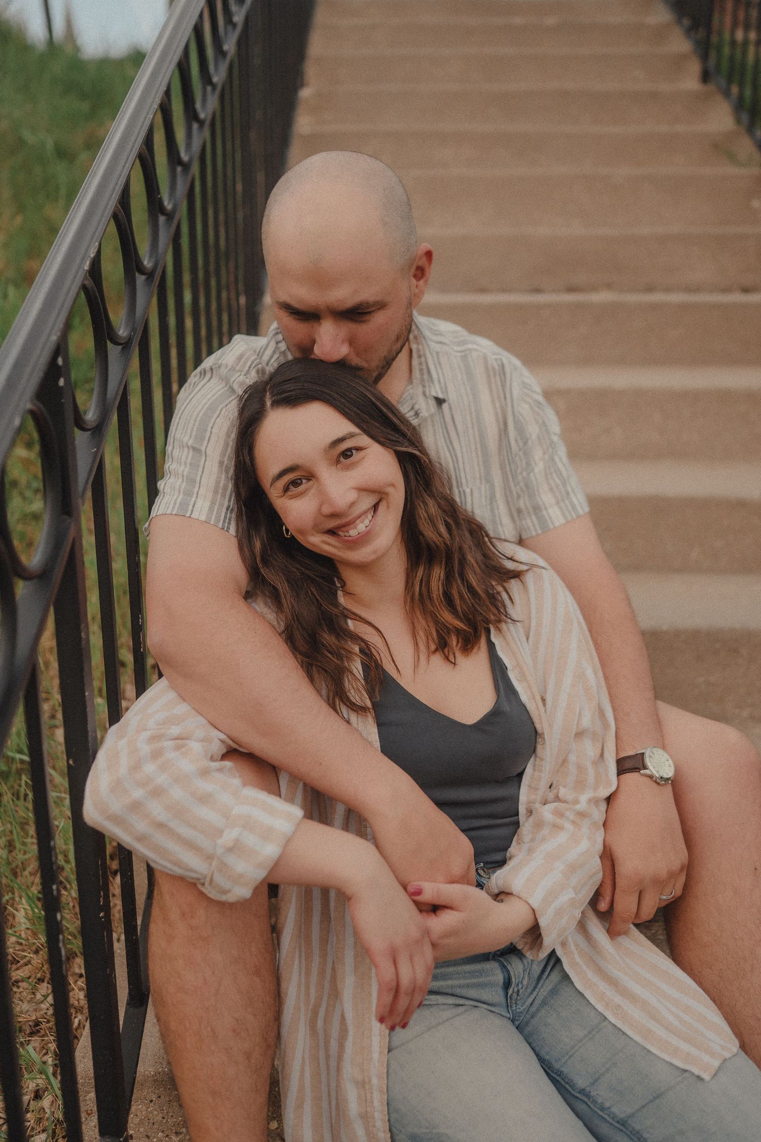 A smiling couple sits on outdoor stairs, the man embracing the woman from behind while she looks at the camera.