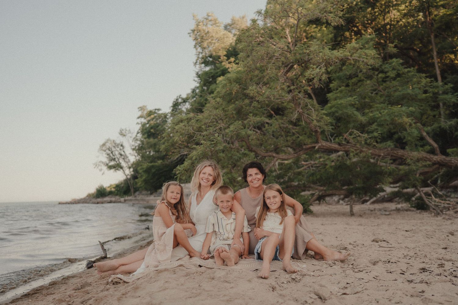 Smiling family of six sitting together on sandy beach with green trees at sunset.