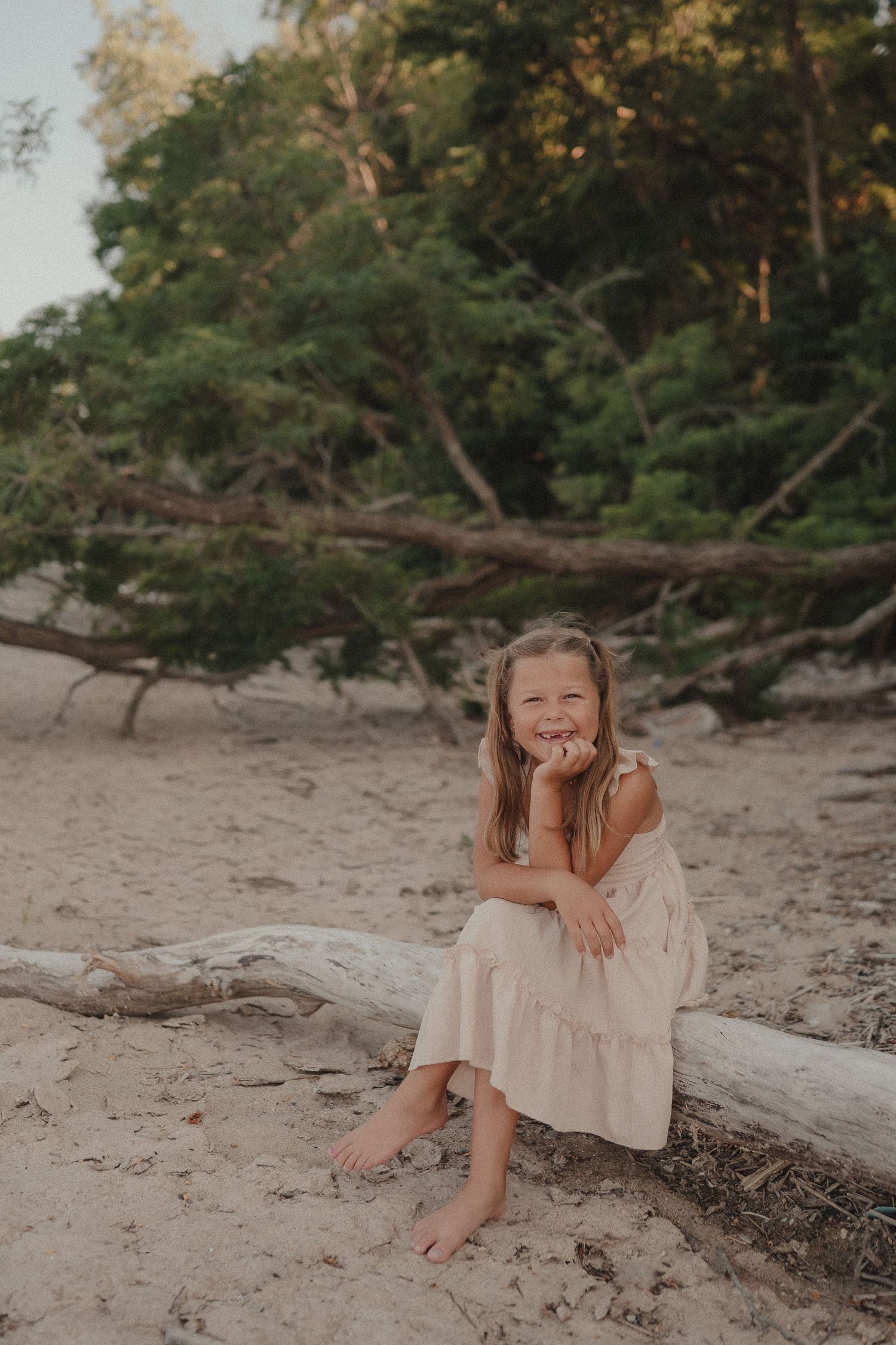 A young girl in a light pink dress sits barefoot on driftwood, smiling on a sandy beach with lush green trees behind her.