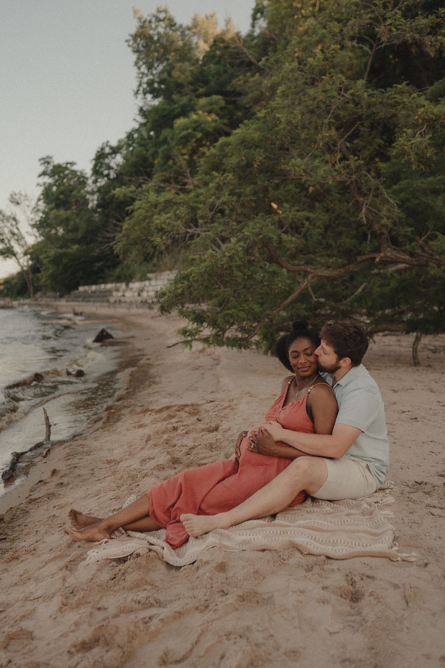 Interracial couple sitting on sandy beach at sunset, man kissing pregnant woman in coral dress near lush green trees.