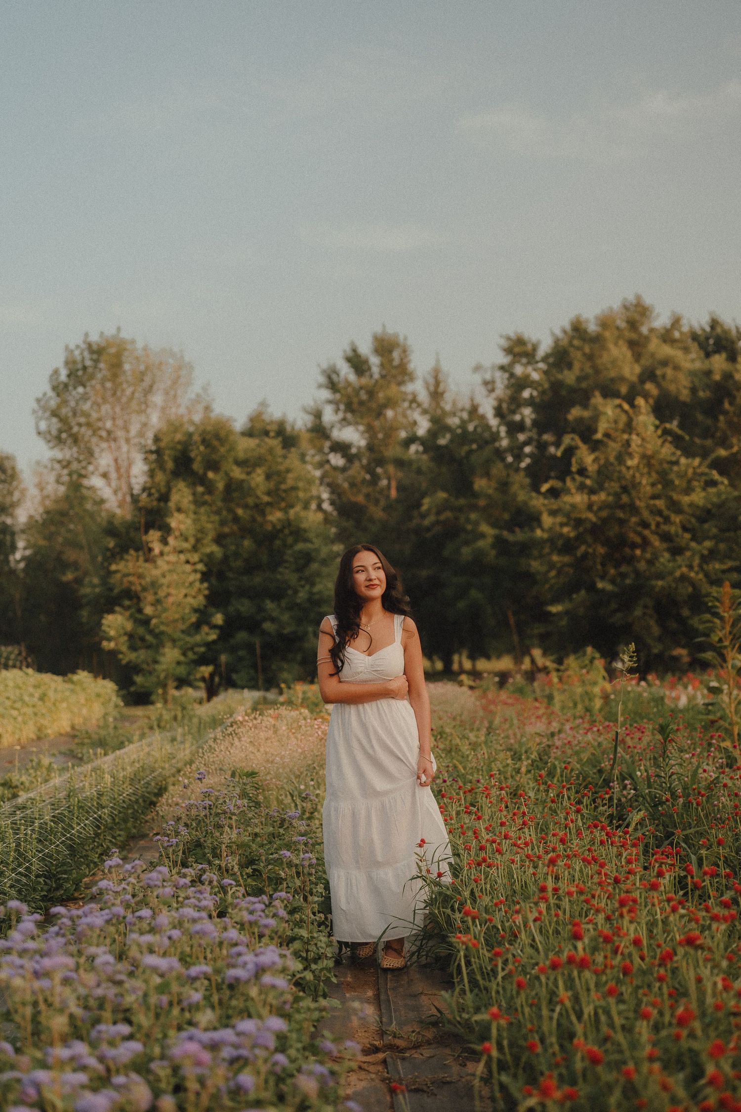 Young woman in white dress standing among colorful wildflowers in a garden path at golden hour.