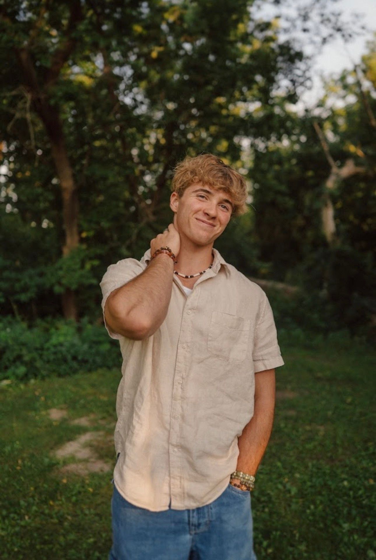 A casual summer portrait in a wooded setting featuring a beige short-sleeve button down shirt and relaxed pose.