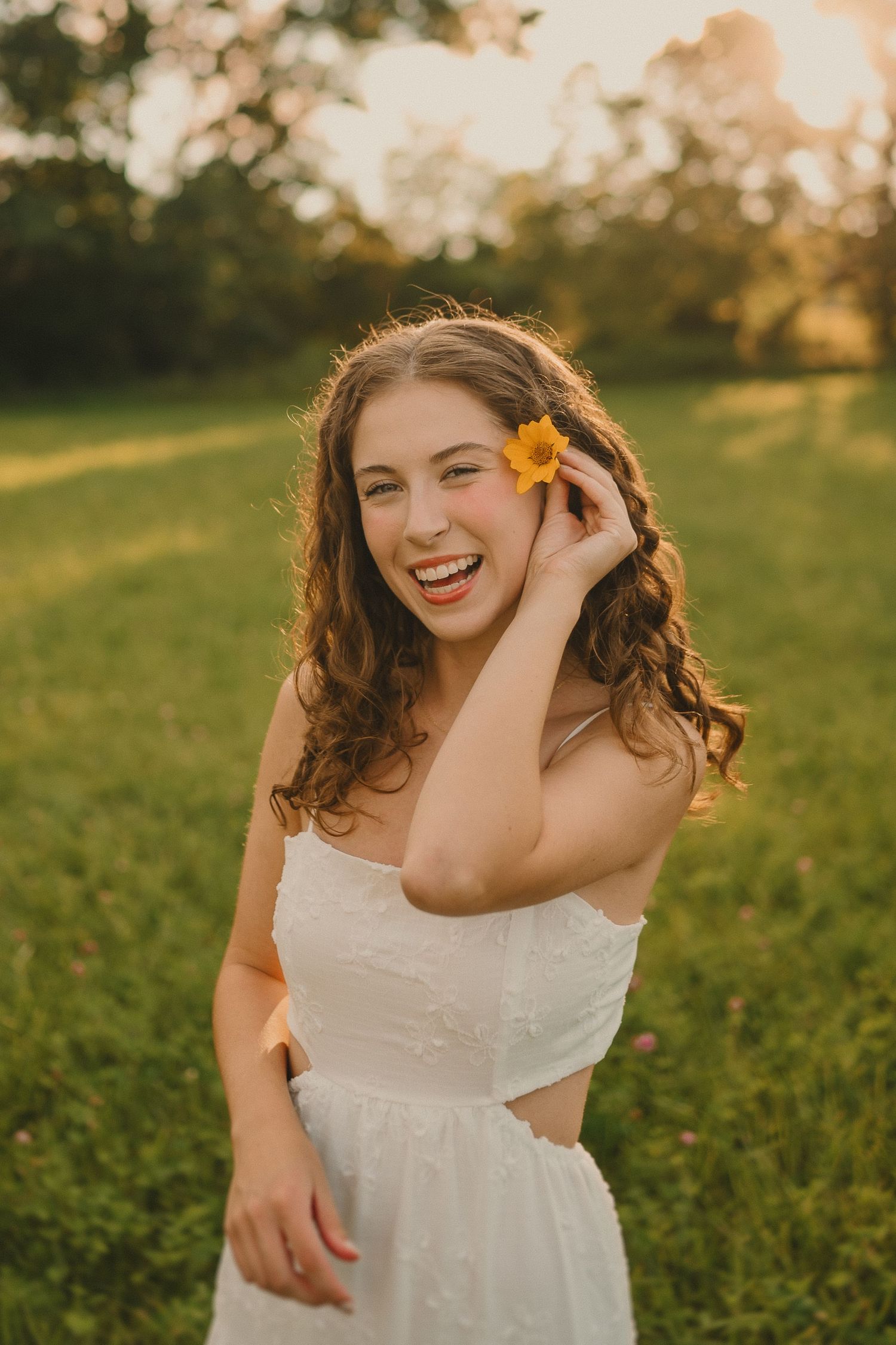 A joyful outdoor portrait features someone in white placing a yellow flower in their curly hair at sunset.