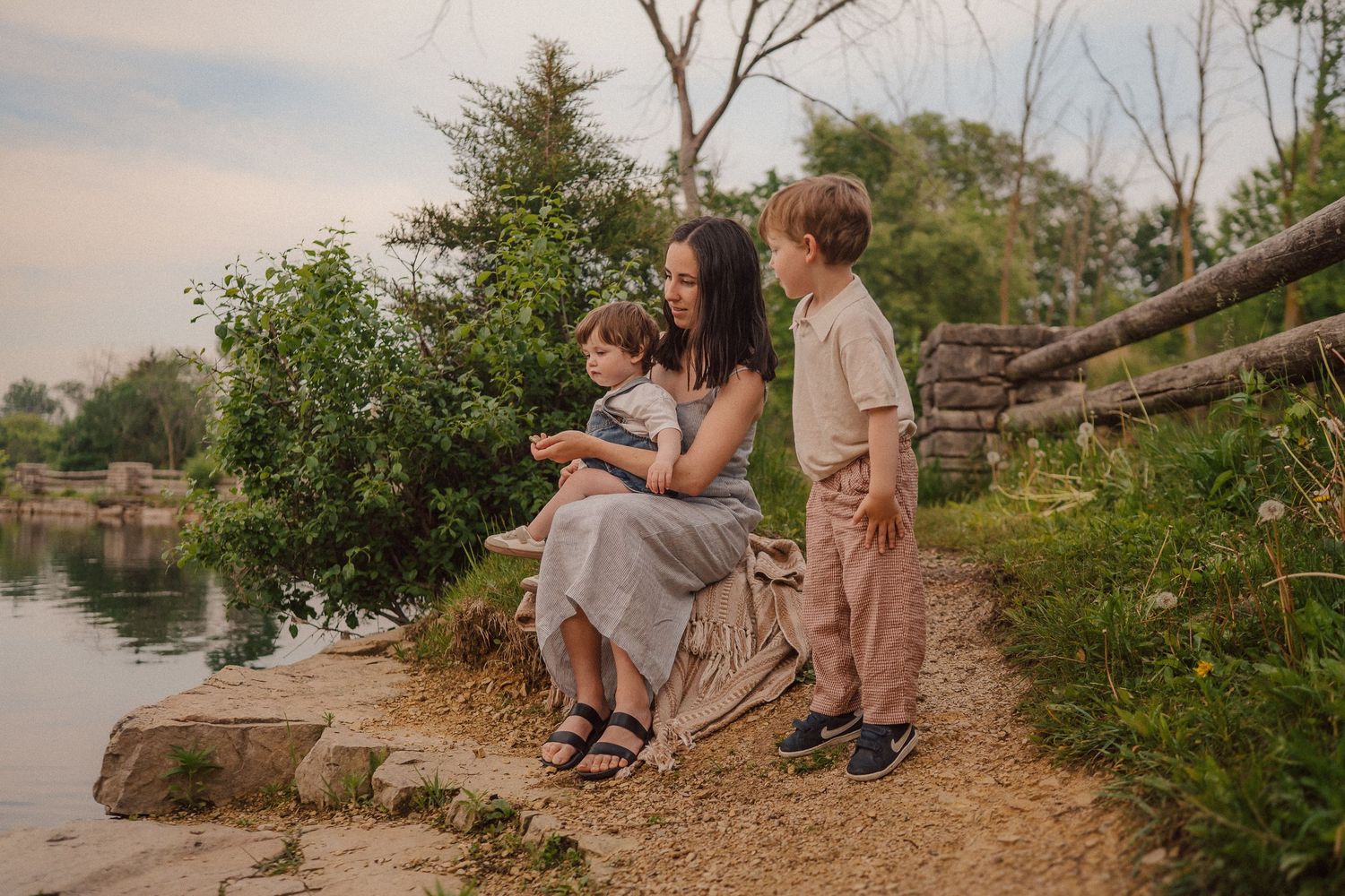 A family scene by a tranquil lake at sunset with trees and rocky shoreline in the background.
