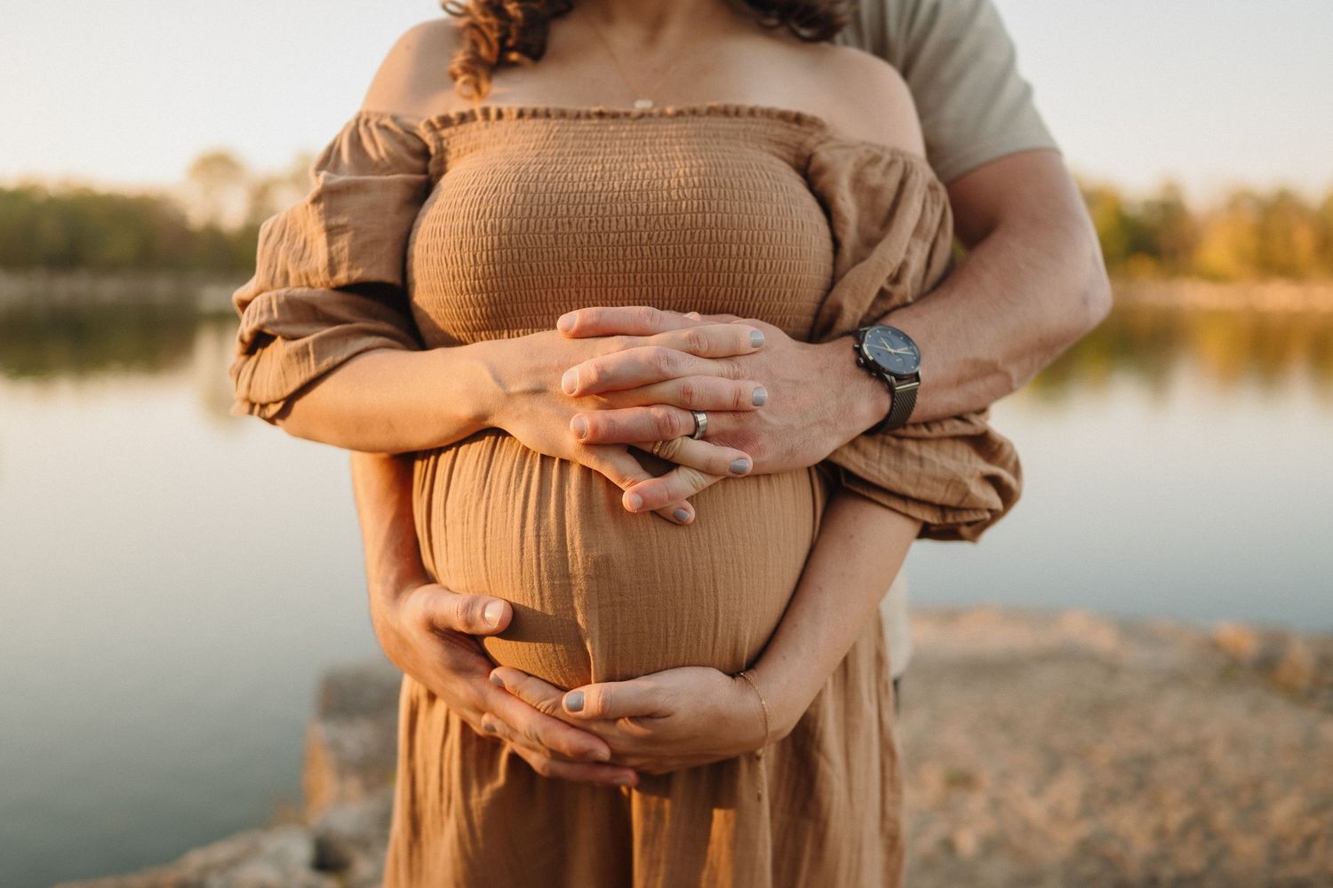 Close-up of hands cradling a pregnant belly in a brown dress against a sunset lake backdrop.