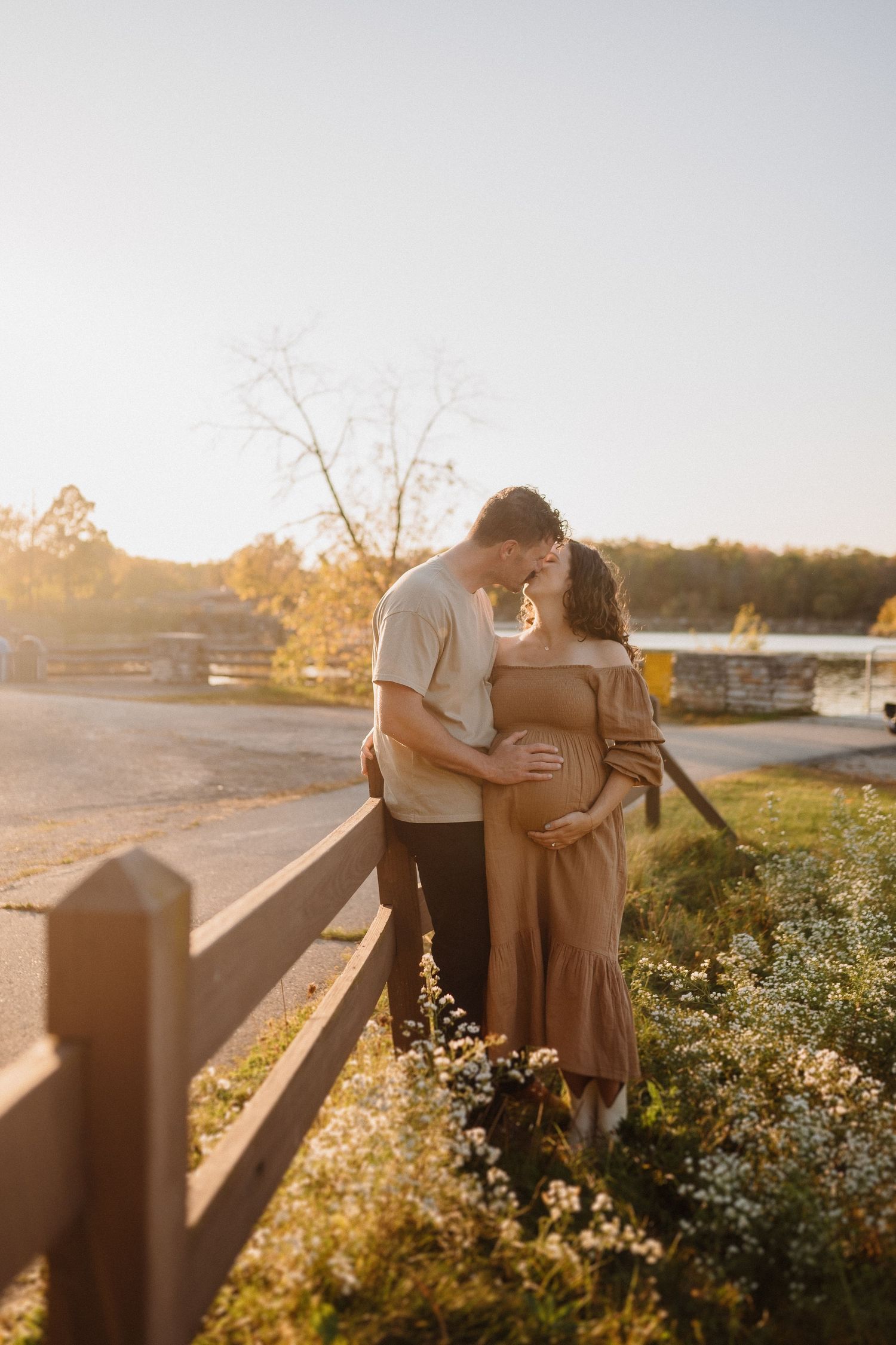 Tender moment between expecting couple by wooden fence at sunset.