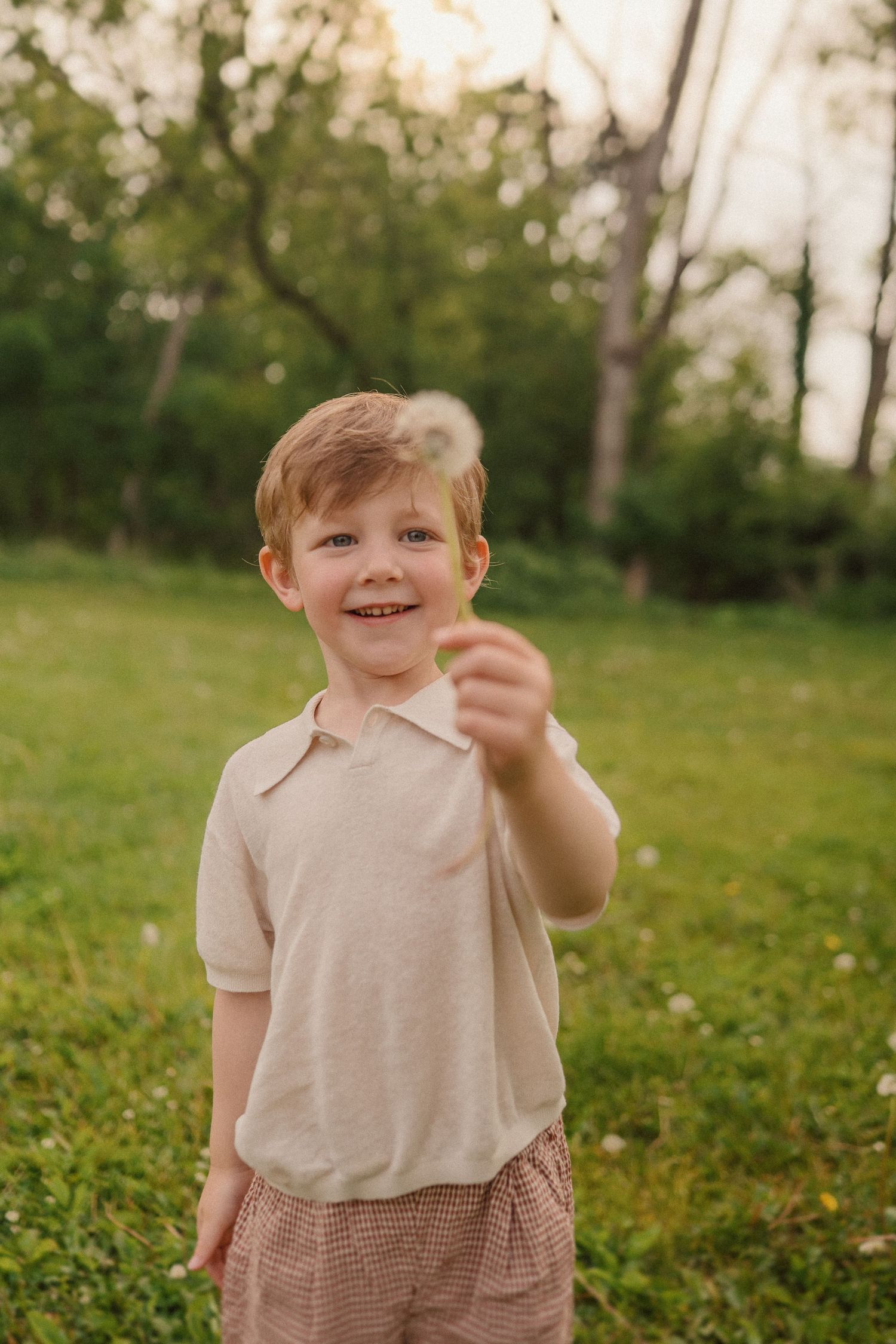 Child in beige polo shirt standing in grassy field at sunset holds something small while smiling.