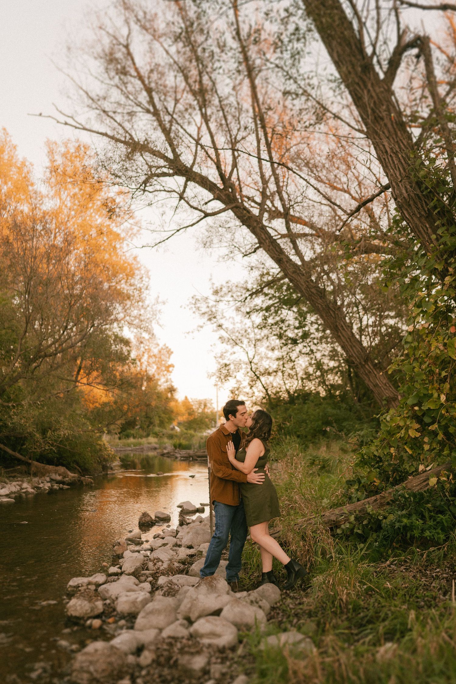 A romantic embrace by the riverside with autumn trees and golden light creating a dreamy atmosphere.