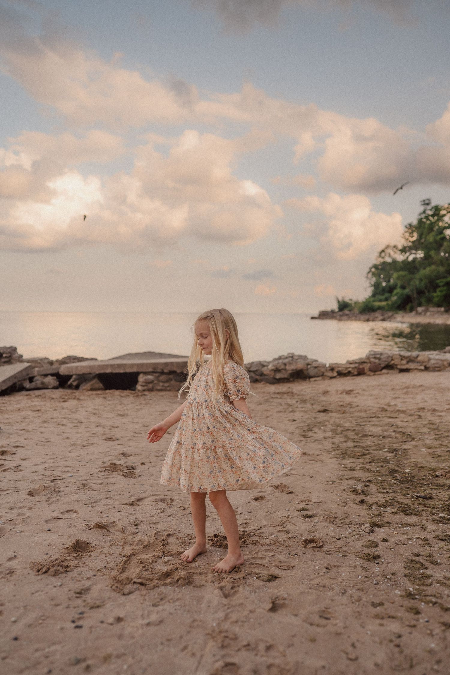 A young girl in a floral dress twirls on the beach at sunset with rocks and trees in the background.