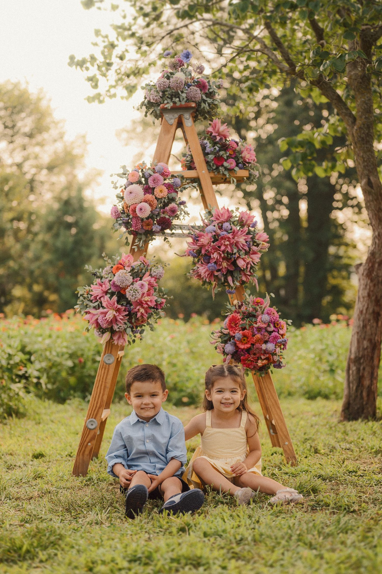 Children sitting on grass near wooden easels decorated with beautiful pink and purple flower arrangements.