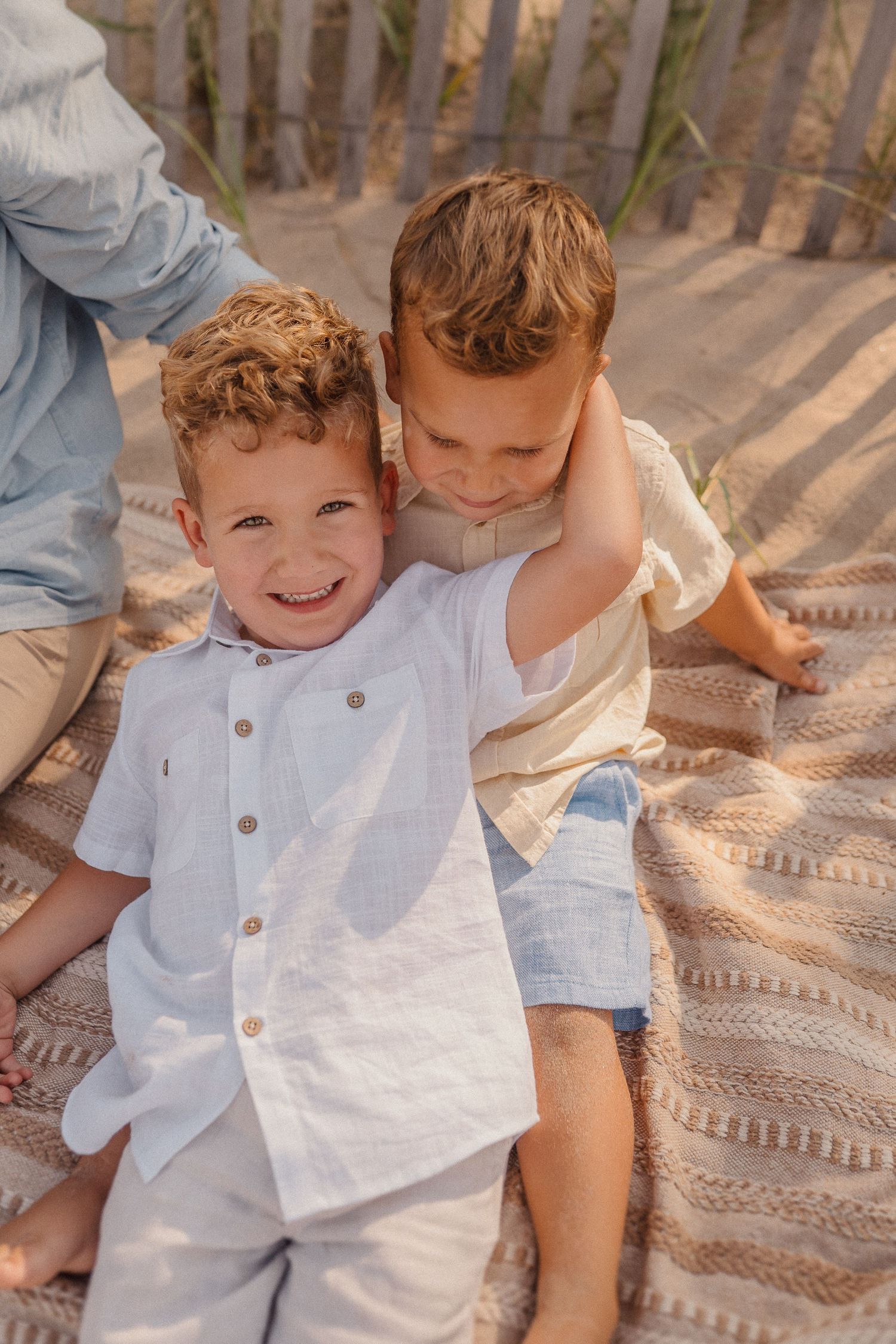 Two young children play together on a blanket by a wooden fence while wearing light summer clothing.