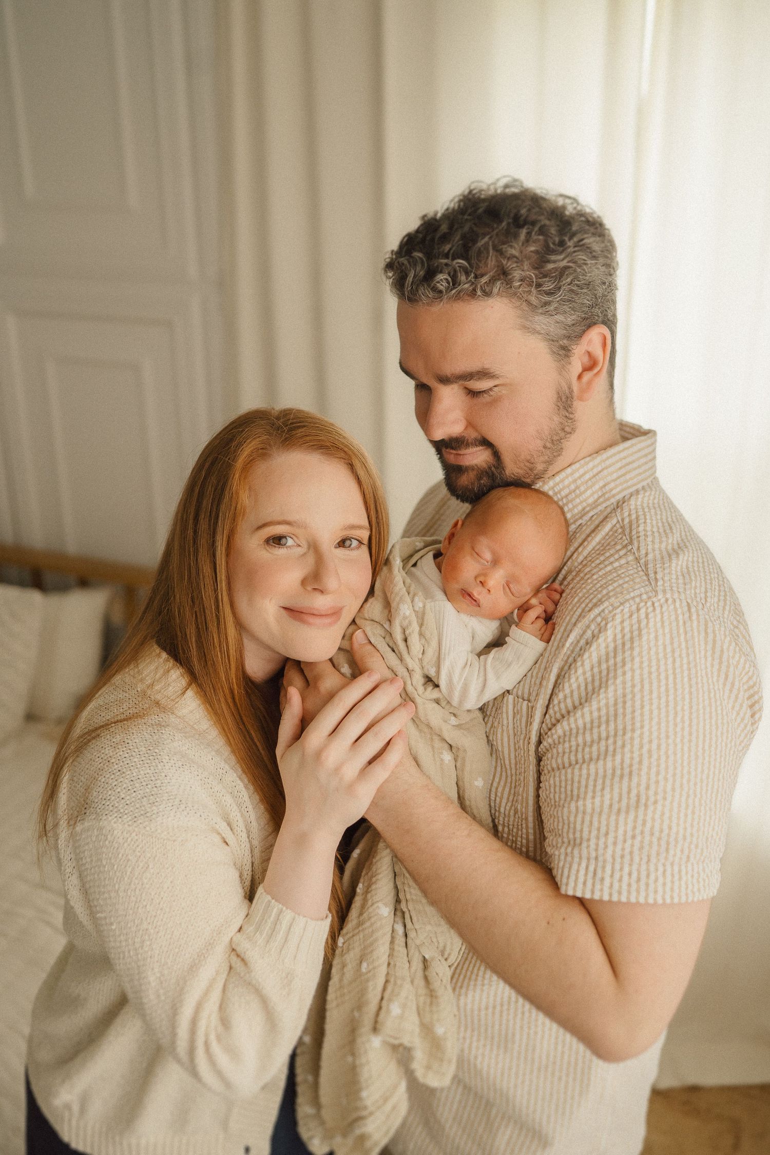 A tender family moment as parents lovingly embrace their sleeping newborn baby in a bright, cozy room.