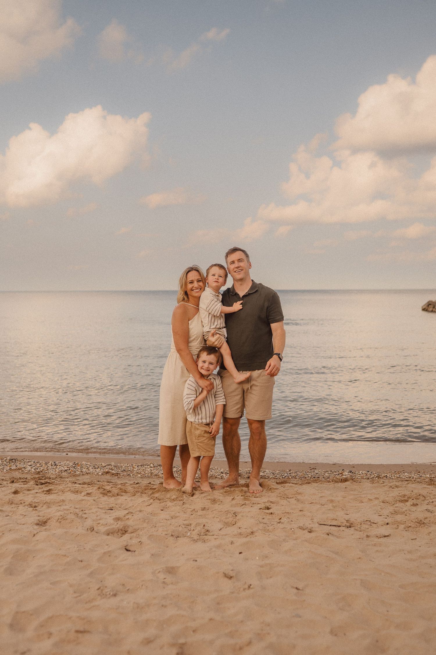 People gathered on a sandy beach at sunset with calm ocean waters and cloudy skies in the background.