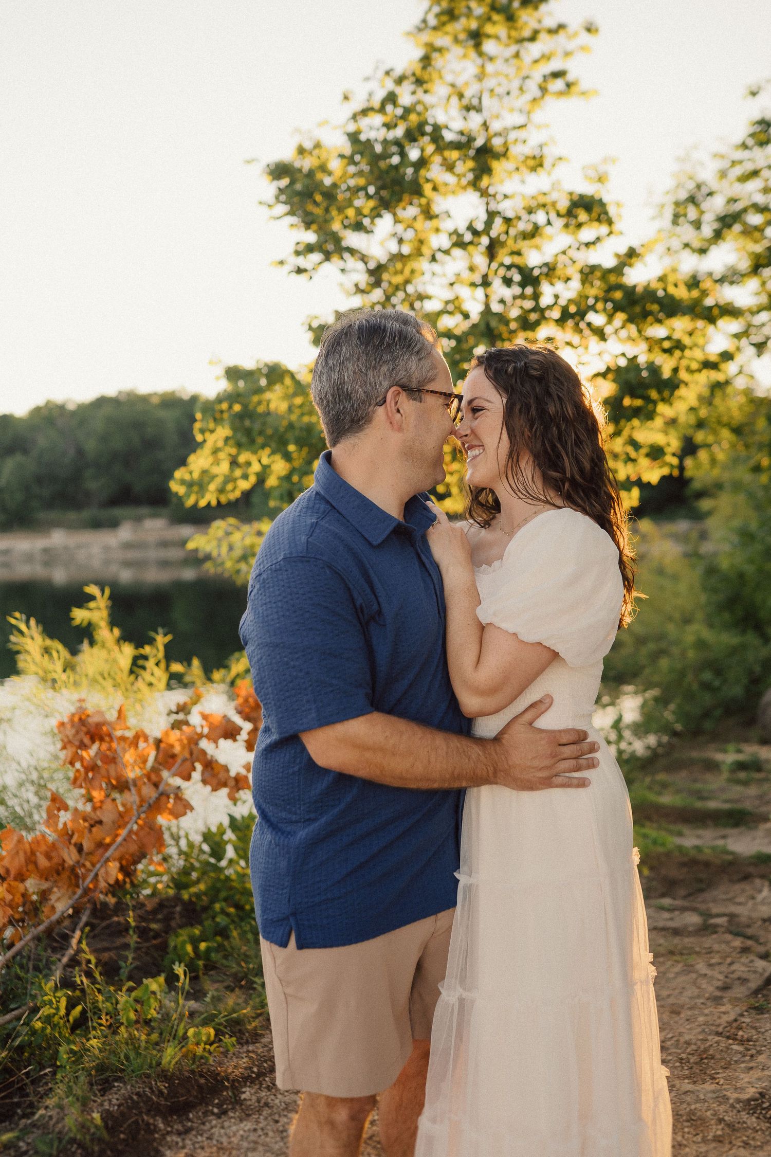 A romantic couple embraces during golden hour sunset in a natural outdoor setting with autumn foliage.