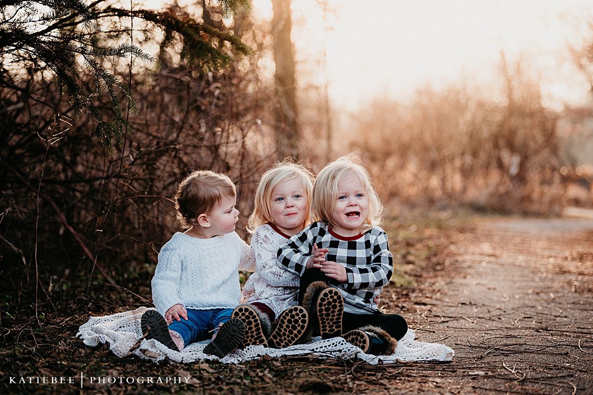 Three small children sit together on a forest path during golden hour.