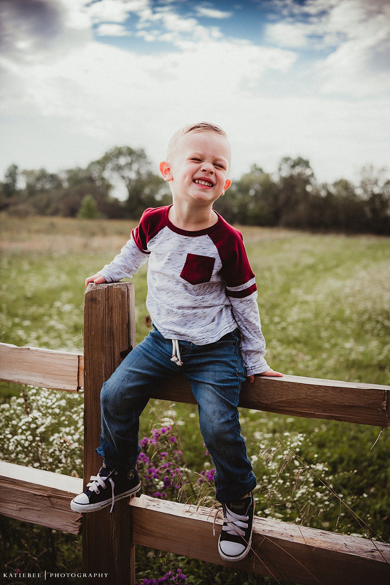 Child sitting on wooden fence post in casual clothing against wildflower field.