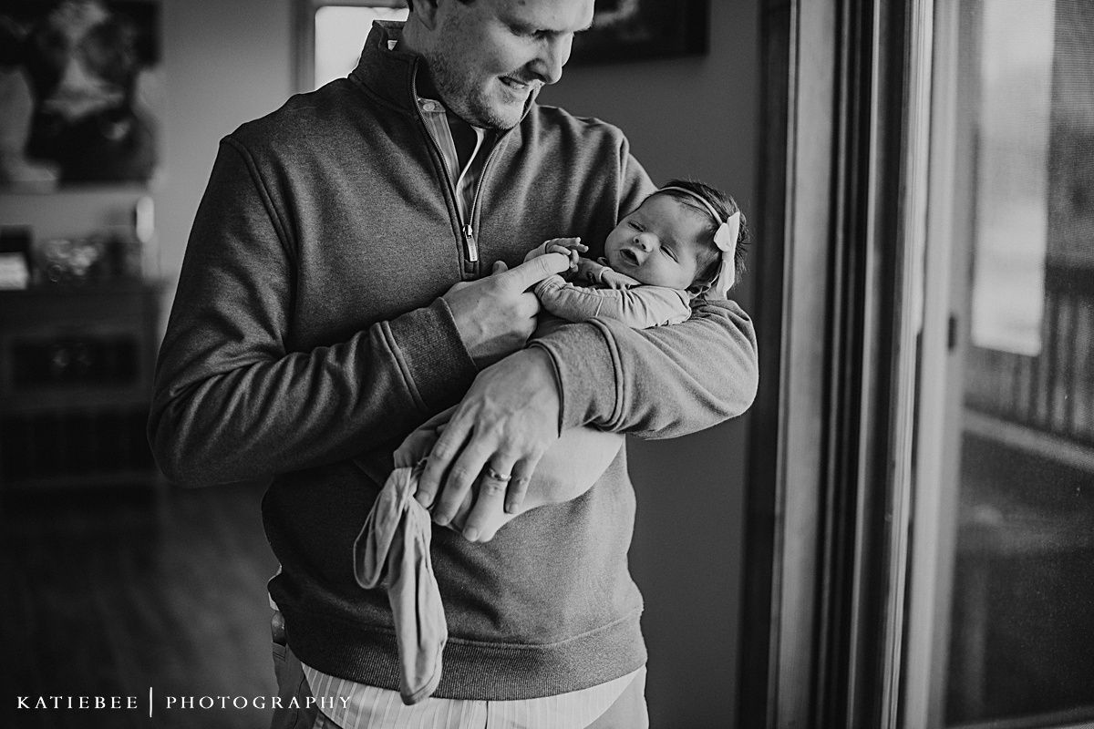 Black and white portrait of parent holding infant near window during lifestyle photo session.