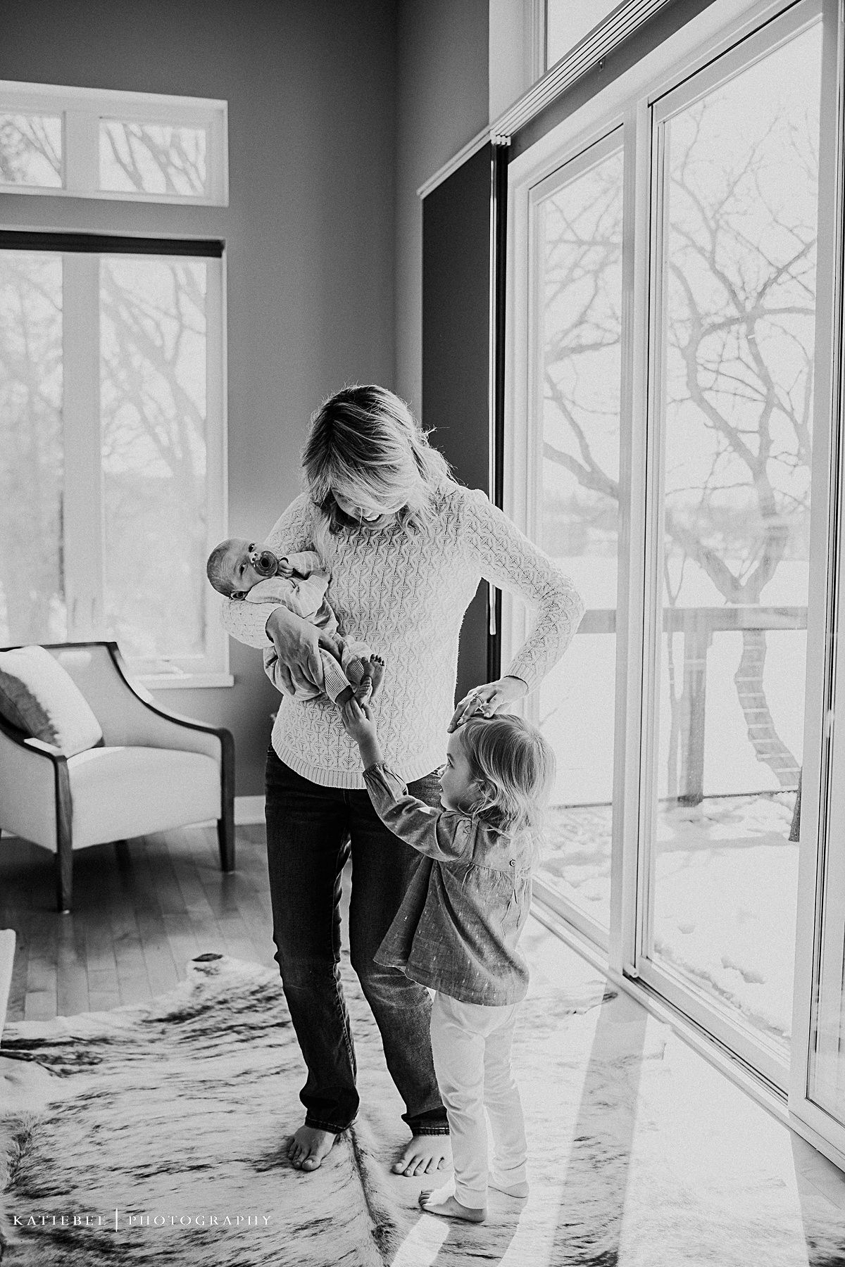 Family shares sweet moment by large windows with newborn and toddler.