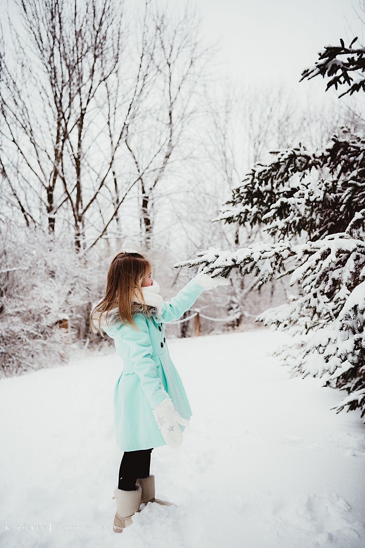 Person in turquoise coat reaching toward snow covered trees.