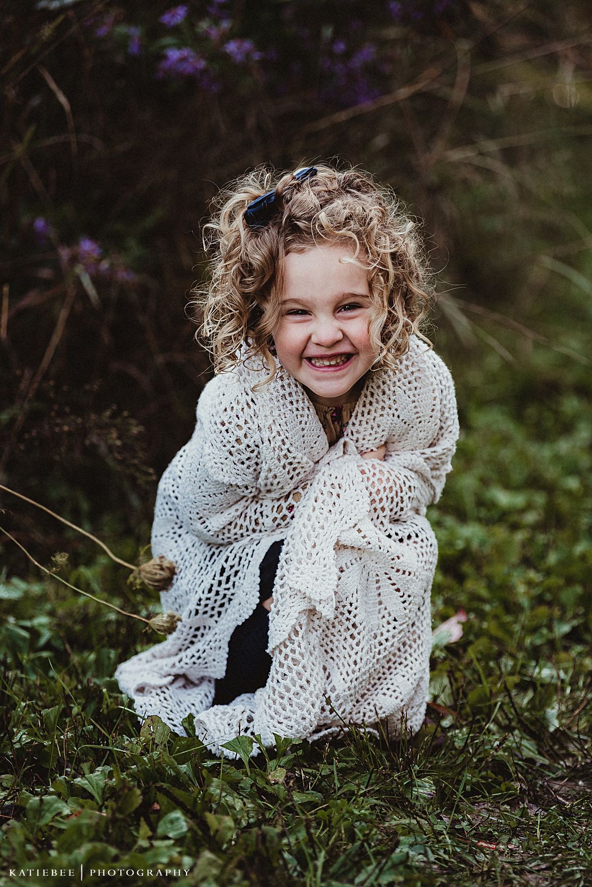 A child wearing a white lace shawl laughs joyfully in a garden setting.