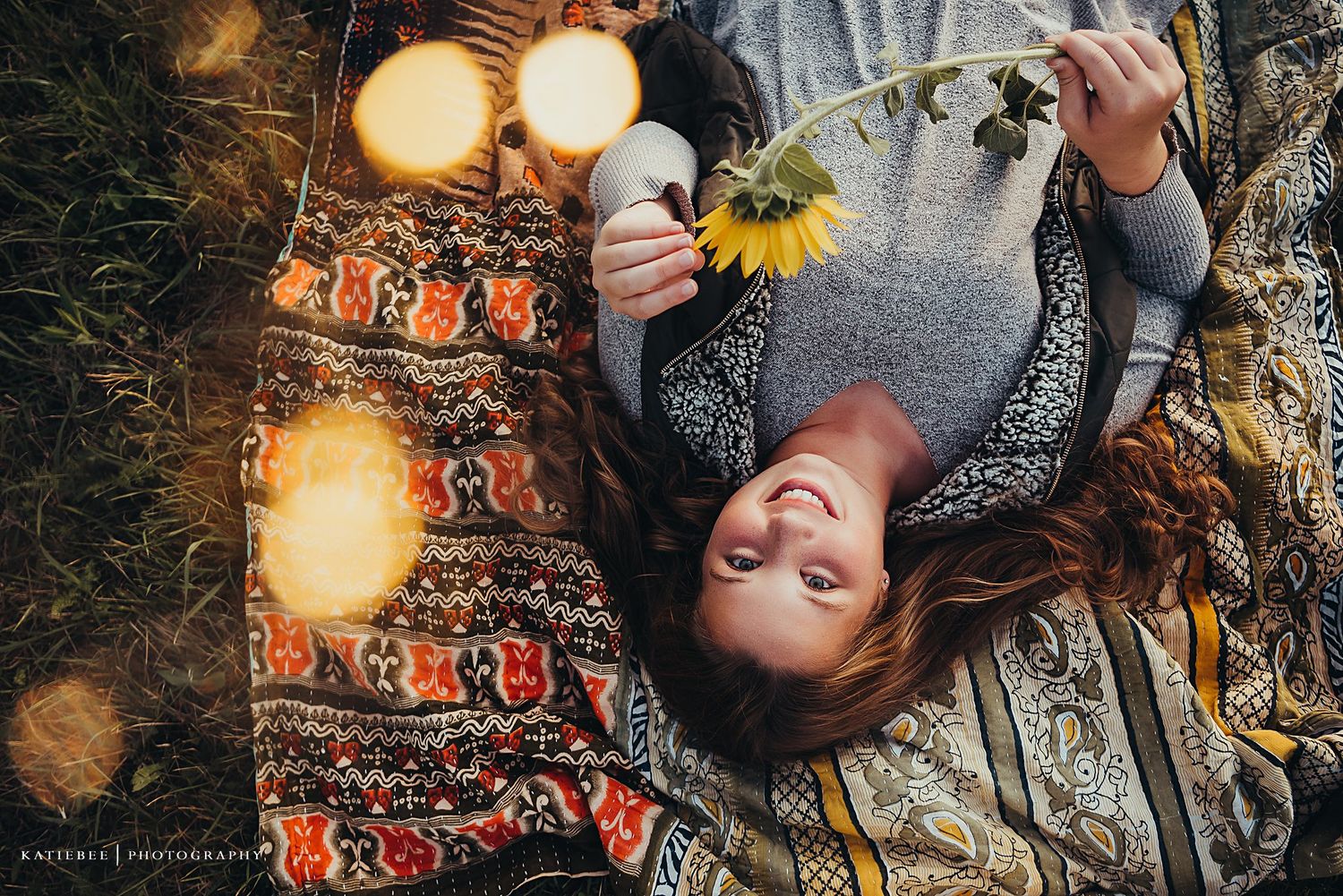 Artistic overhead portrait on a patterned blanket with string lights and sunflower.