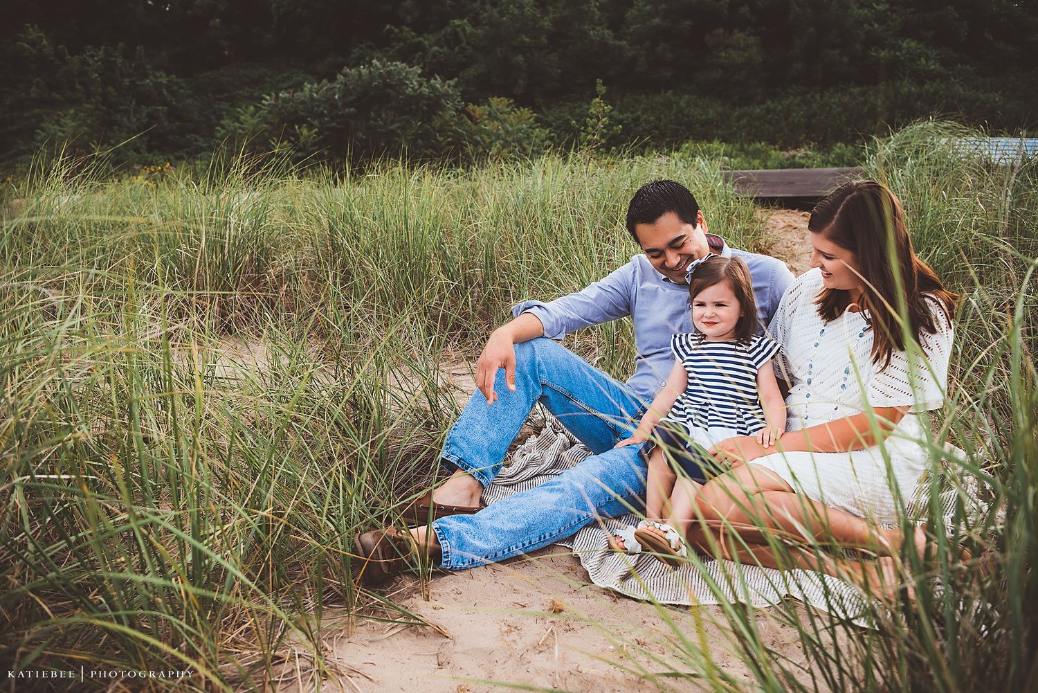 A family shares joyful laughter while sitting together on a blanket in tall grass near the beach.