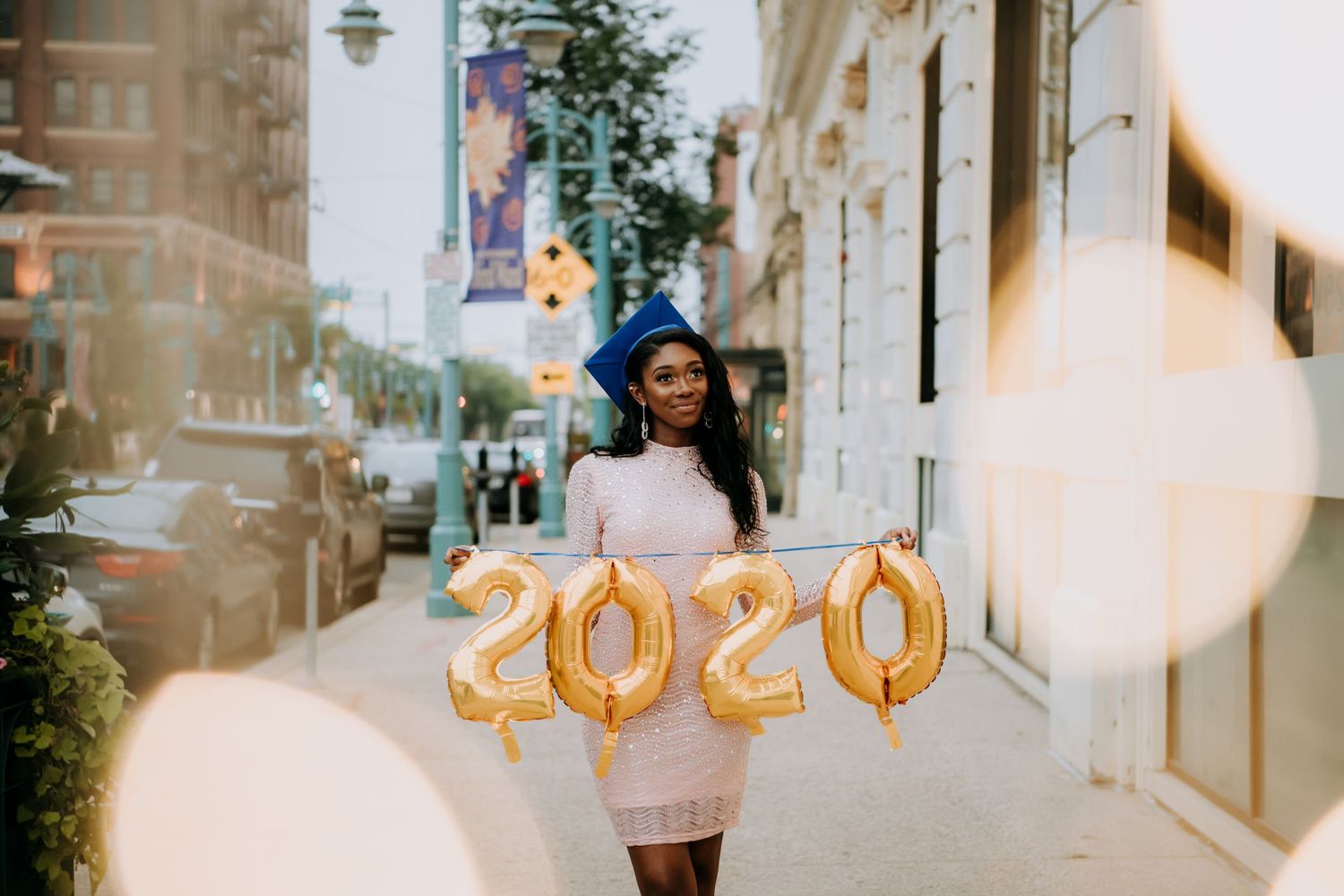 Graduate holding gold 2020 balloons on a city street while wearing a light colored dress and cap.