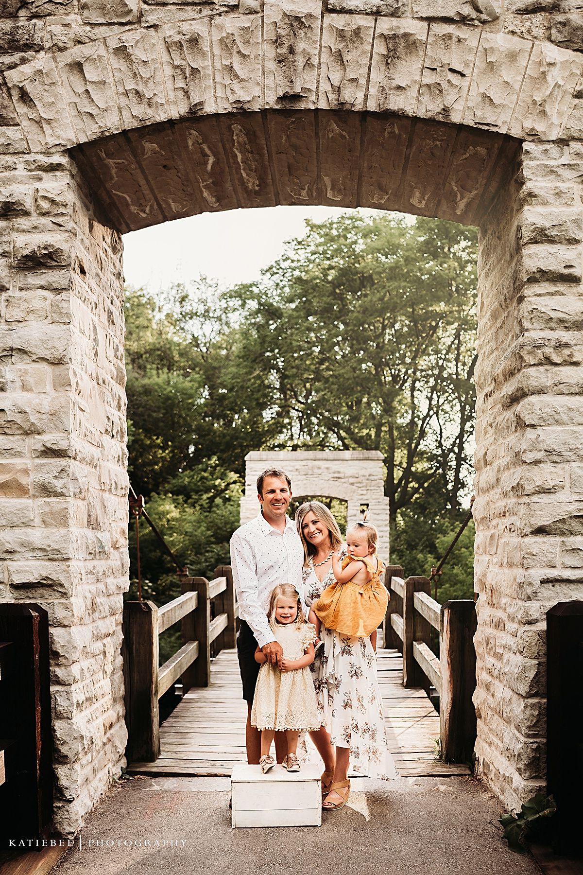 Stone archway frames family portrait on rustic wooden bridge pathway.