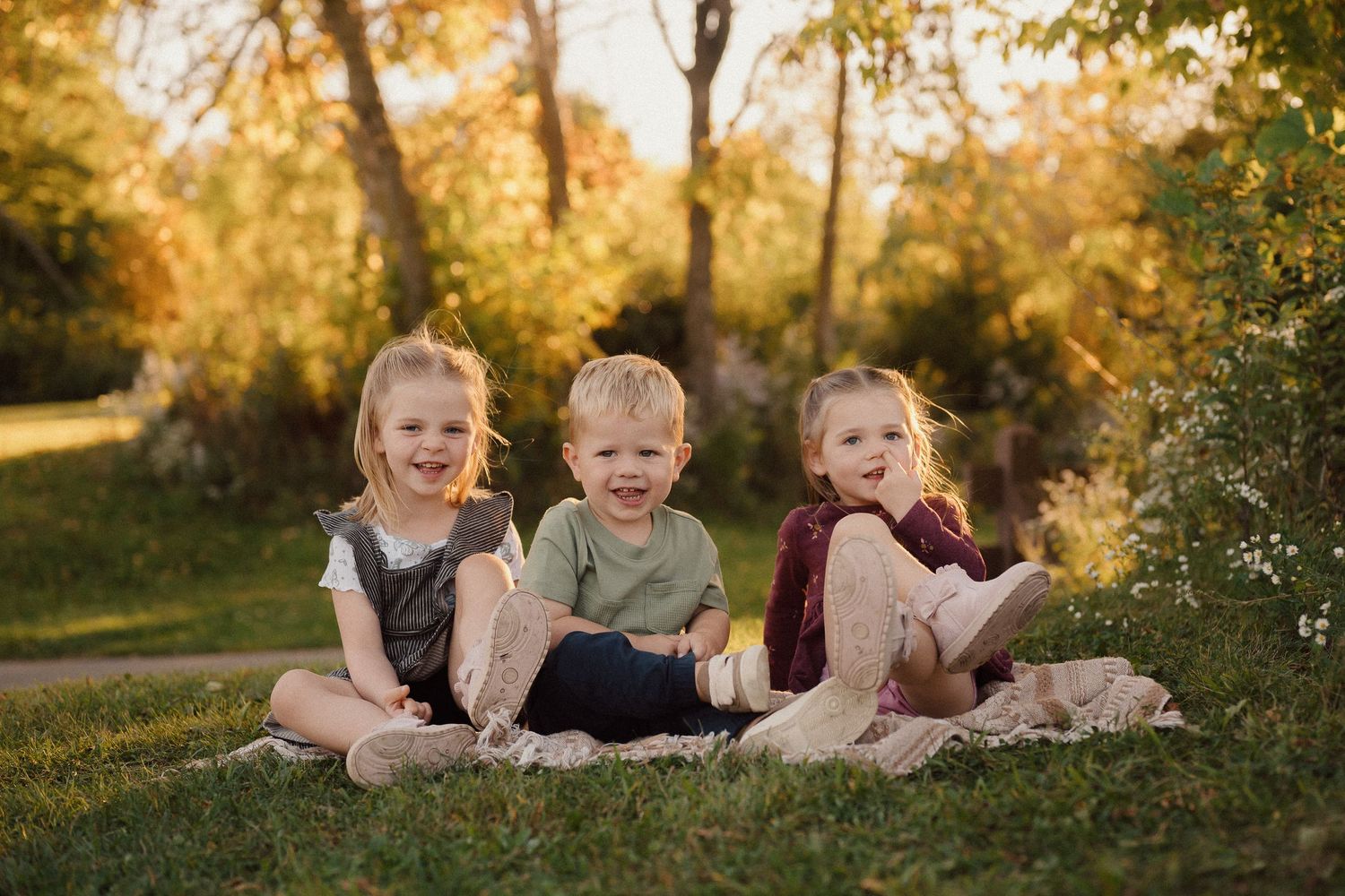 Three children sitting together on grass during a golden autumn afternoon in a park setting.