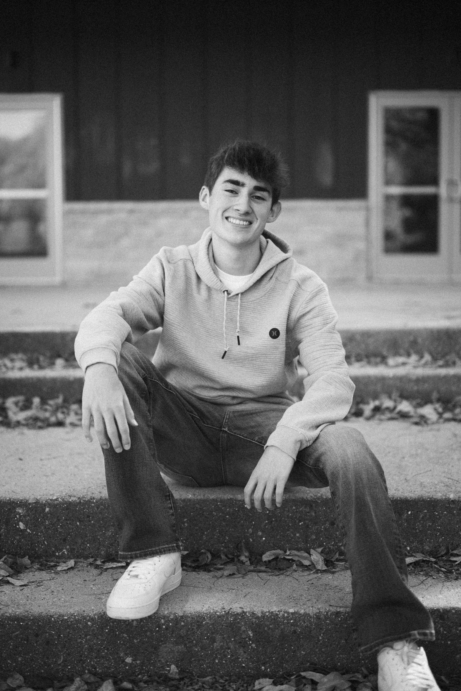 A casual black and white portrait of someone sitting on concrete steps wearing a hoodie and jeans.