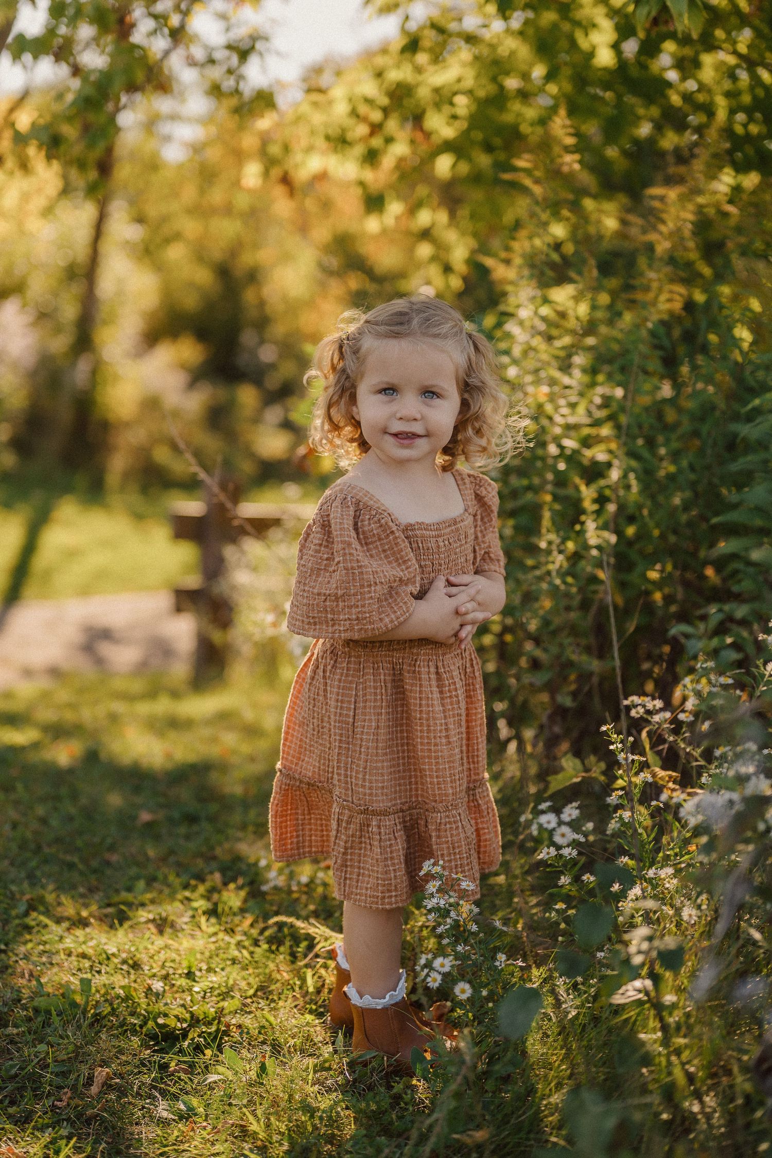 A child in a brown gingham dress plays joyfully in a sunlit garden with lush greenery and flowers.