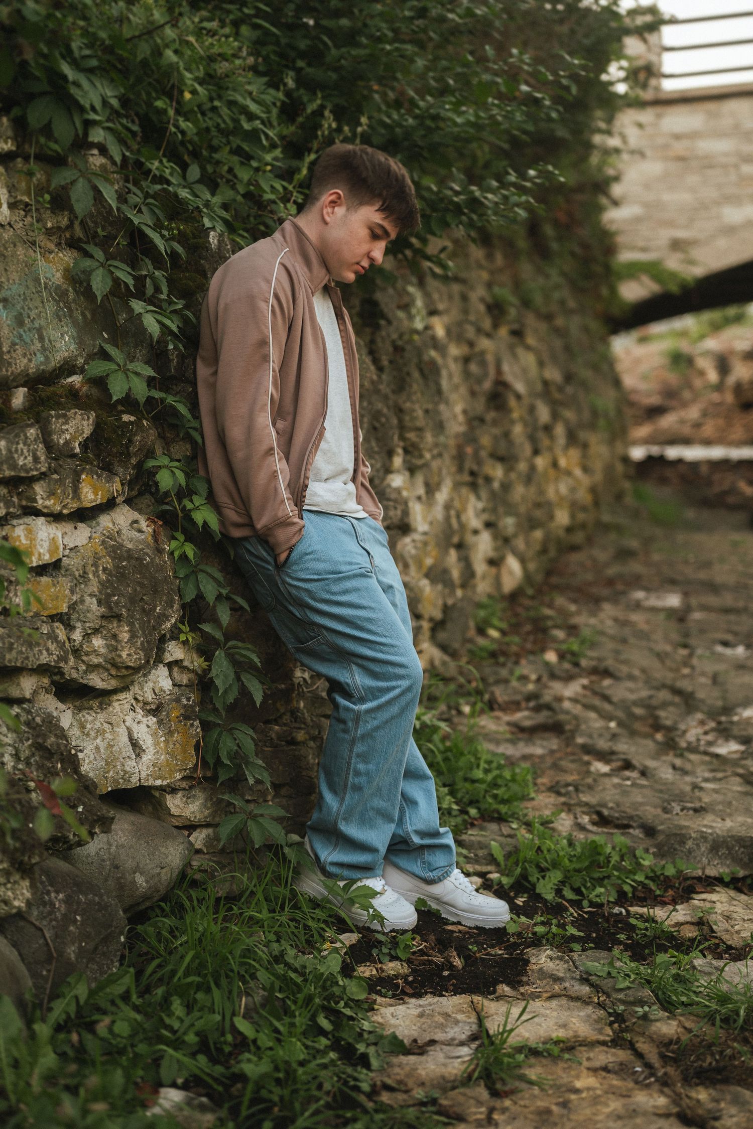 A small child wearing a brown jacket and light blue pants walks along a stone wall path in a garden setting.