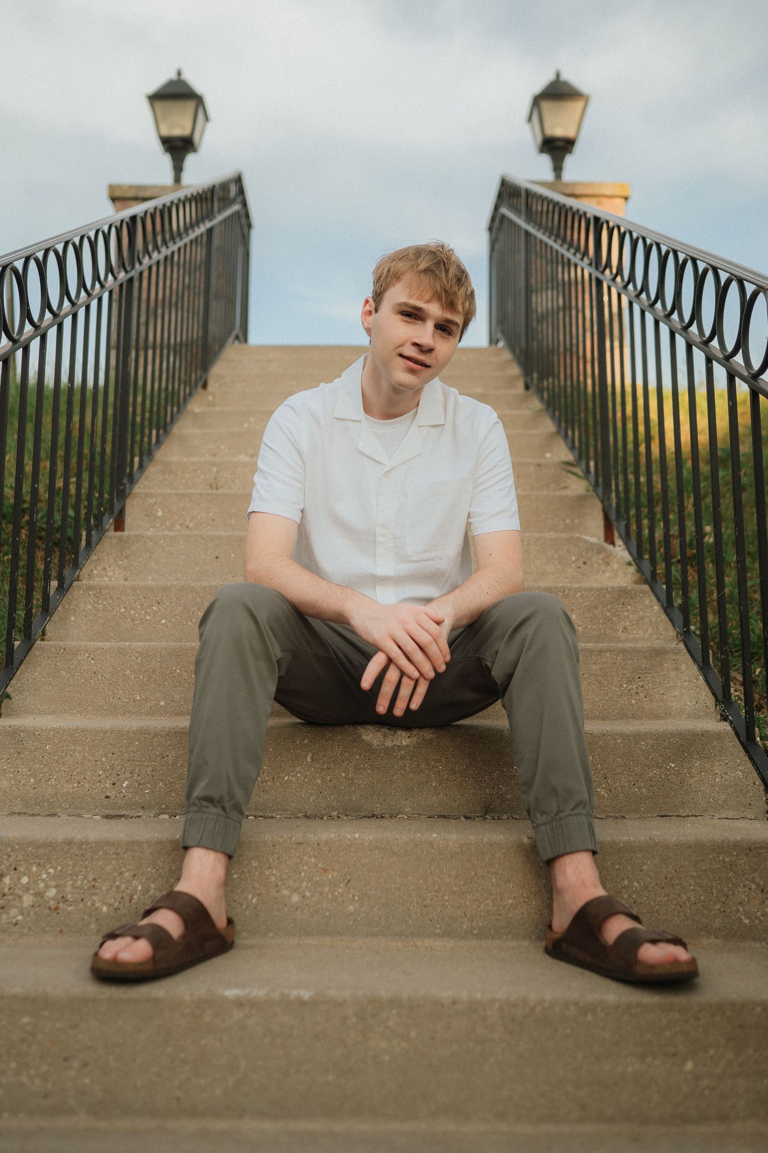 A person in a white t-shirt and olive pants sits on concrete steps between ornate railings and vintage lampposts.