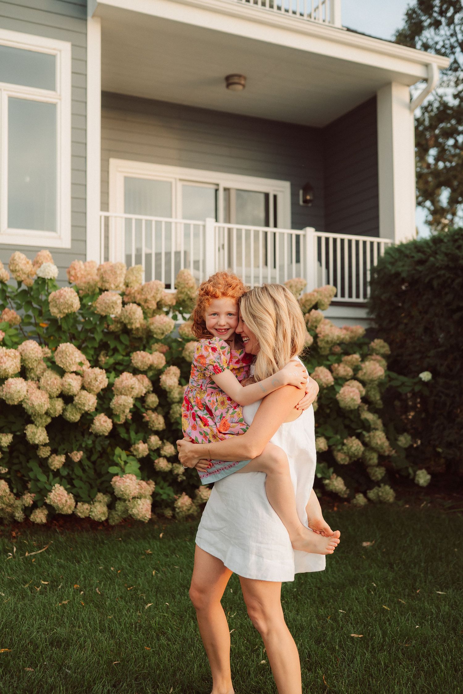 Two people embrace joyfully on a green lawn with blooming hydrangea bushes and white house exterior in the background.