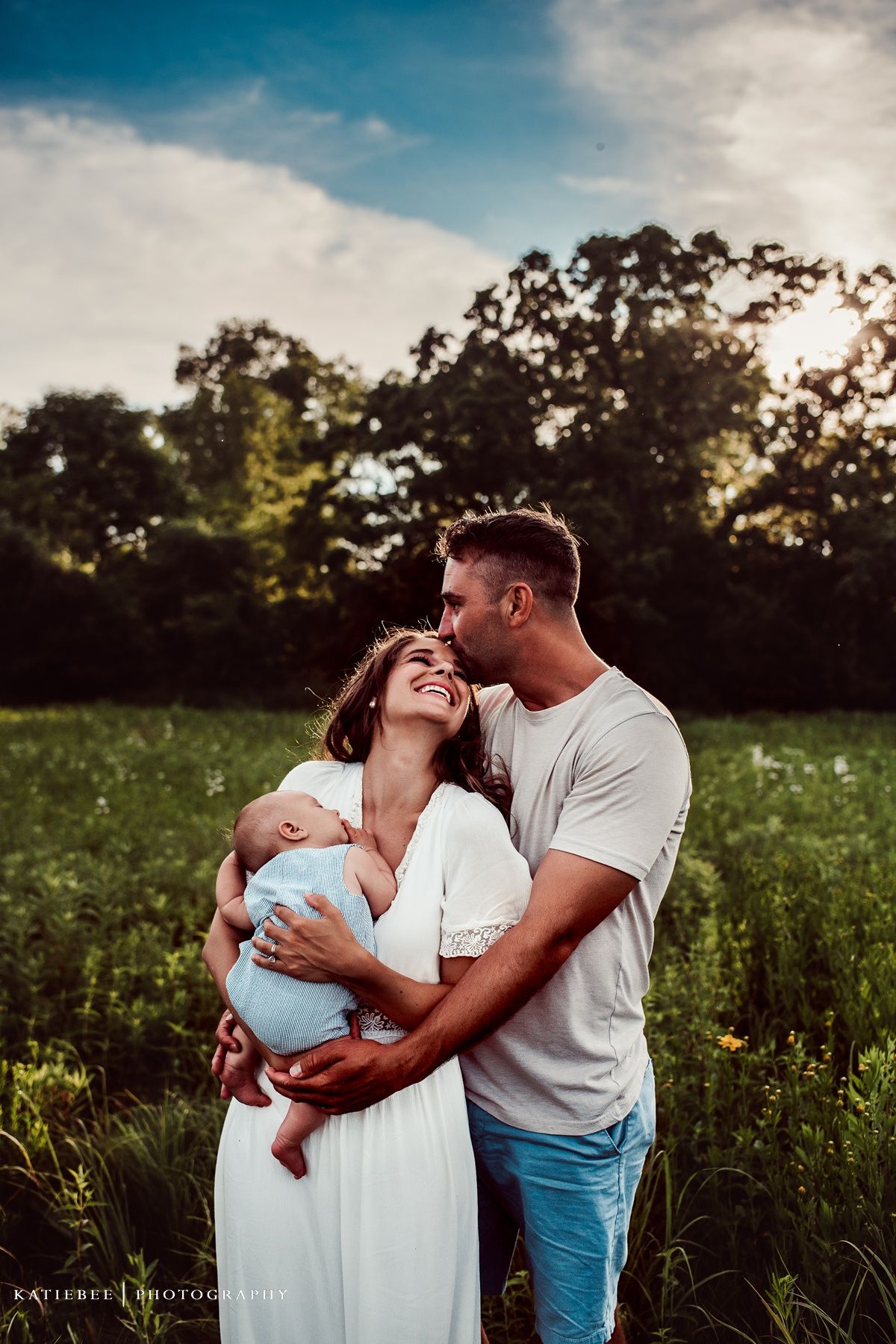 Family moment in field at sunset with couple holding baby in white dress.