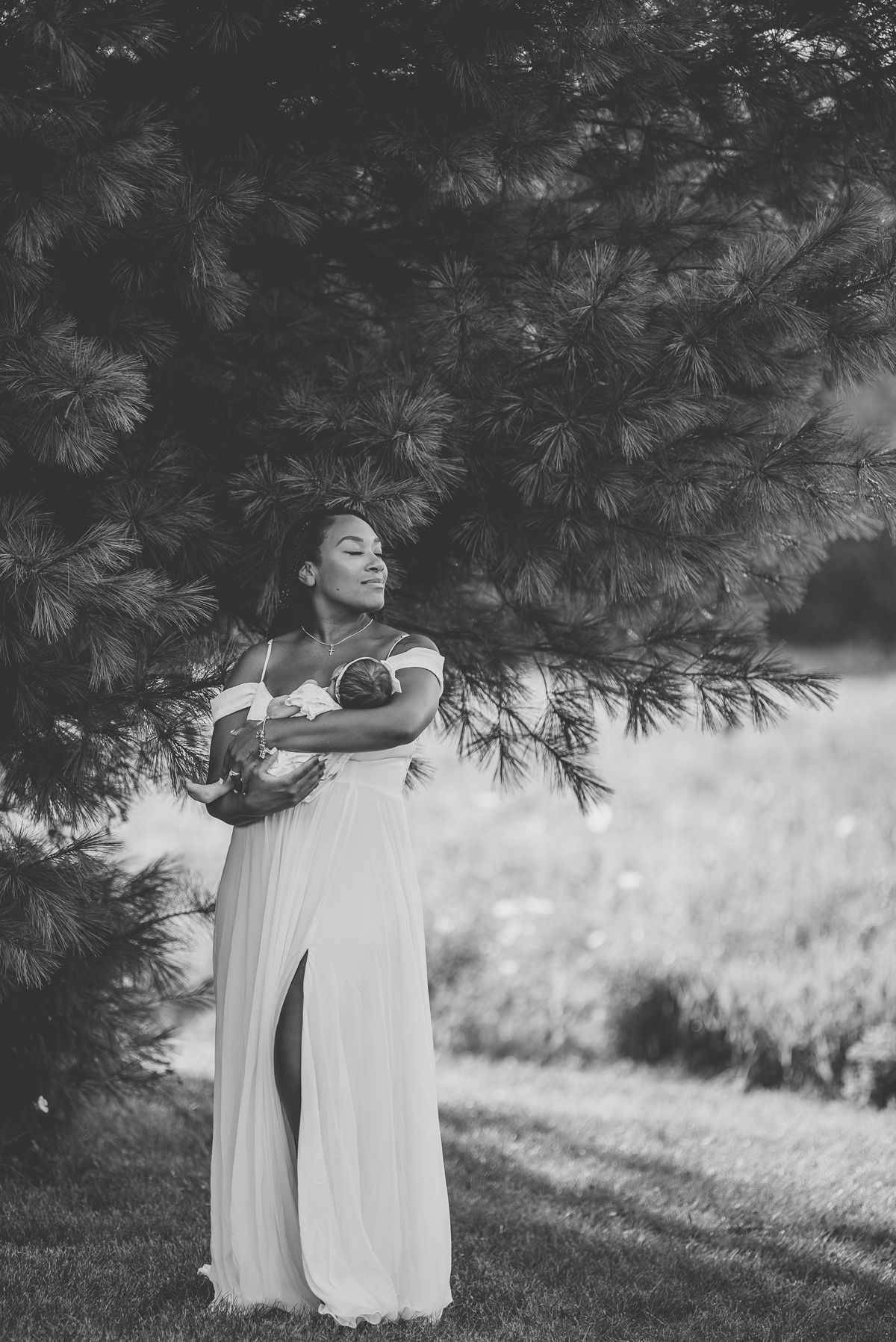 A dreamy black and white portrait of someone in a flowing white dress posed under evergreen branches.