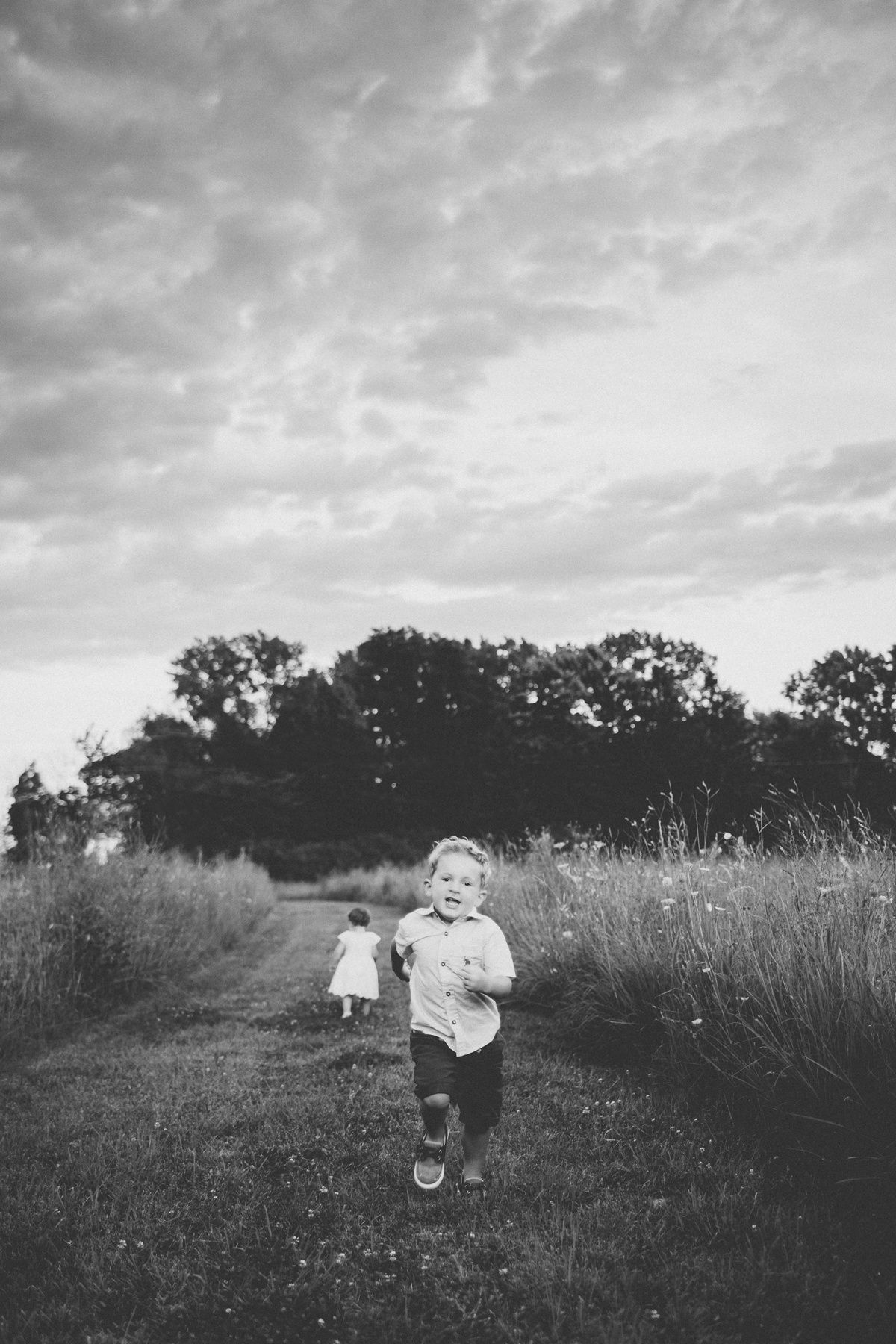 Black and white image of a child running through a rural path with dramatic sky overhead.