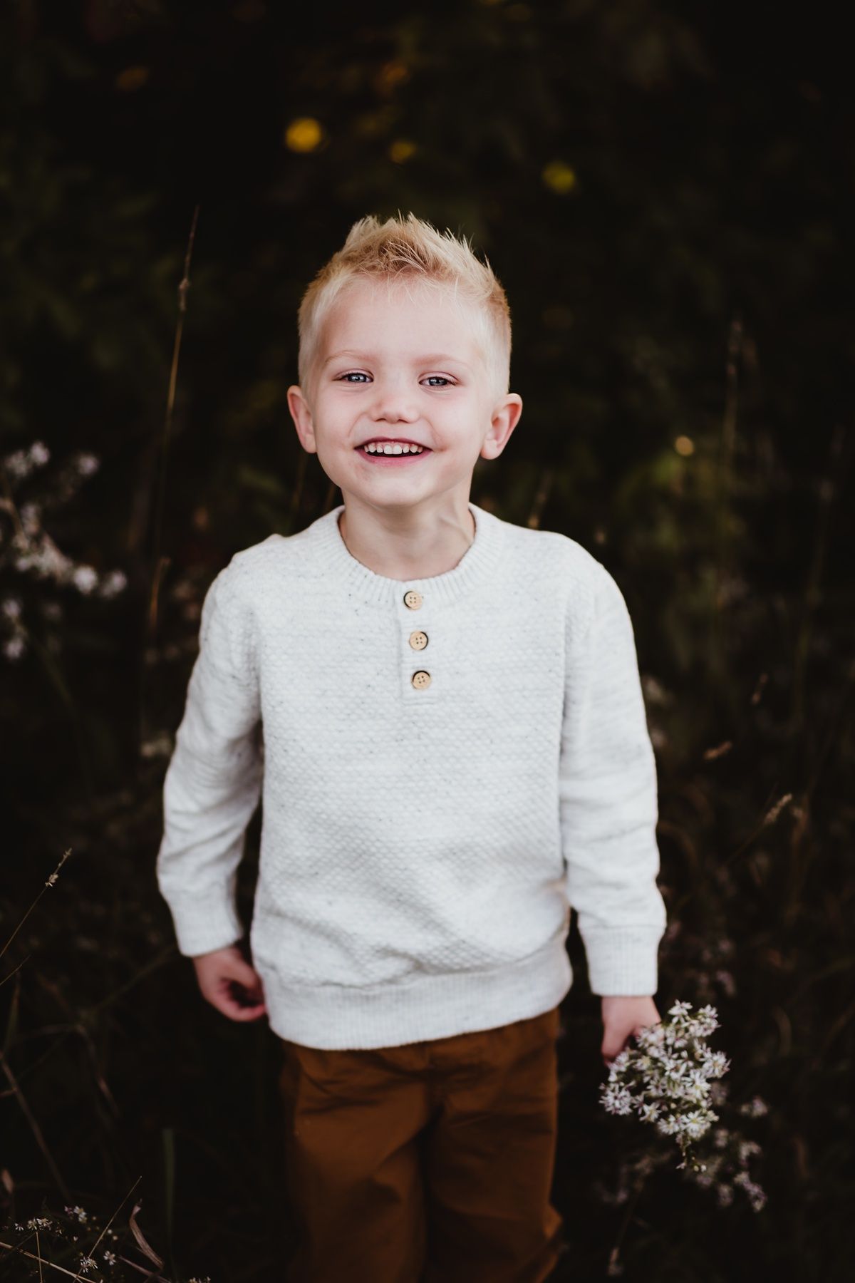 Young child in white knit sweater smiling outdoors against dark background.