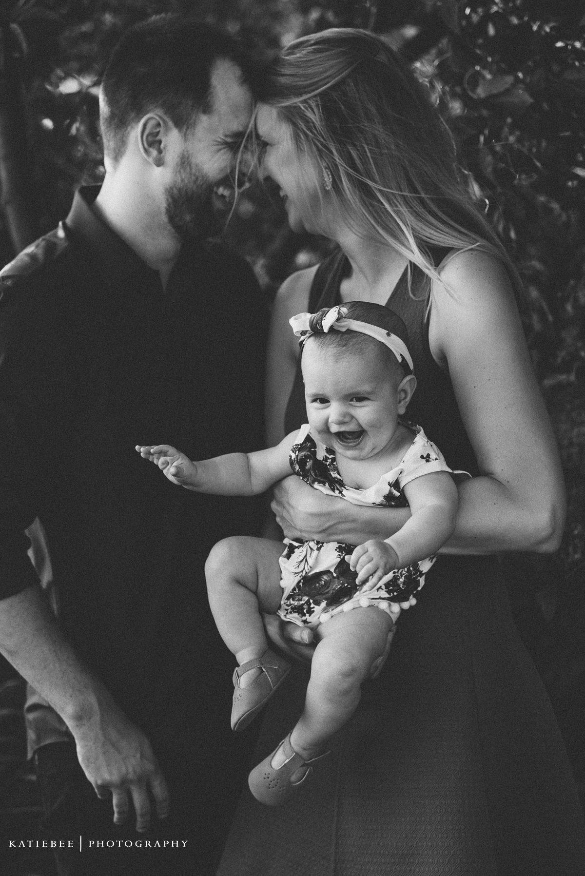 Black and white portrait captures an intimate family moment with a smiling baby in floral outfit.